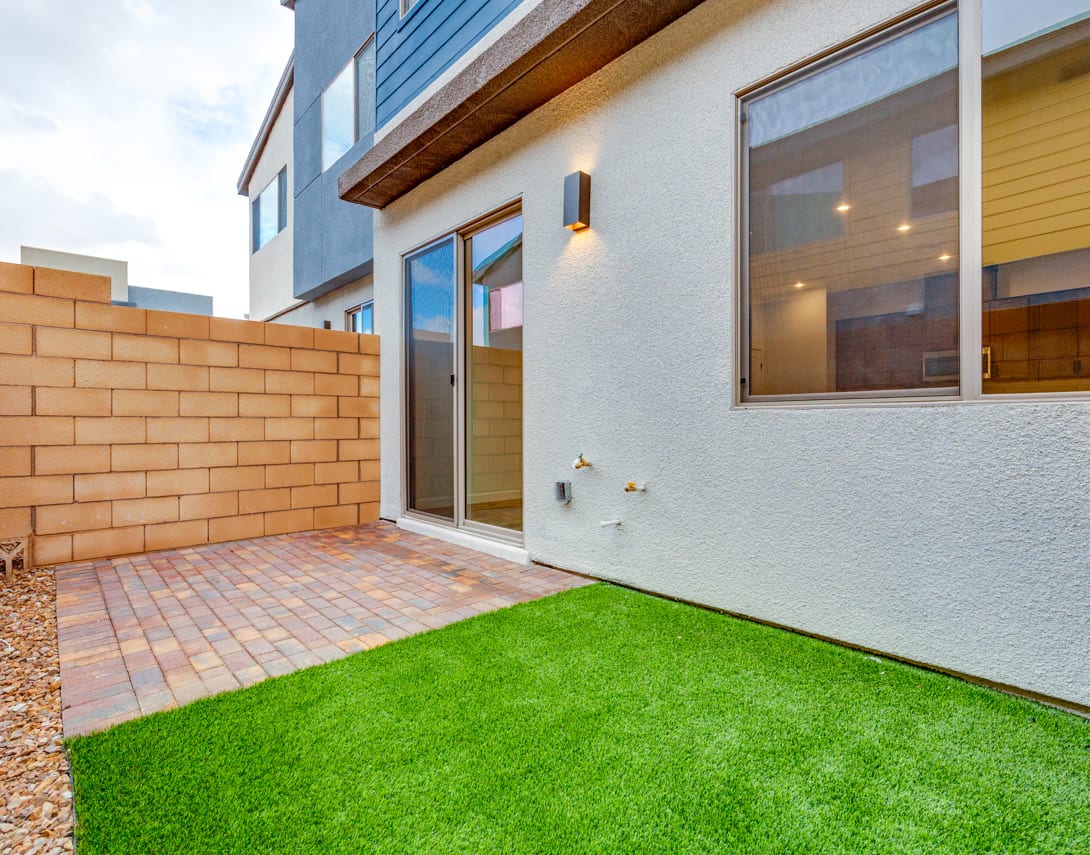 A modern, two-story residential building with a brick wall and a small patio area covered in artificial grass in the foreground.