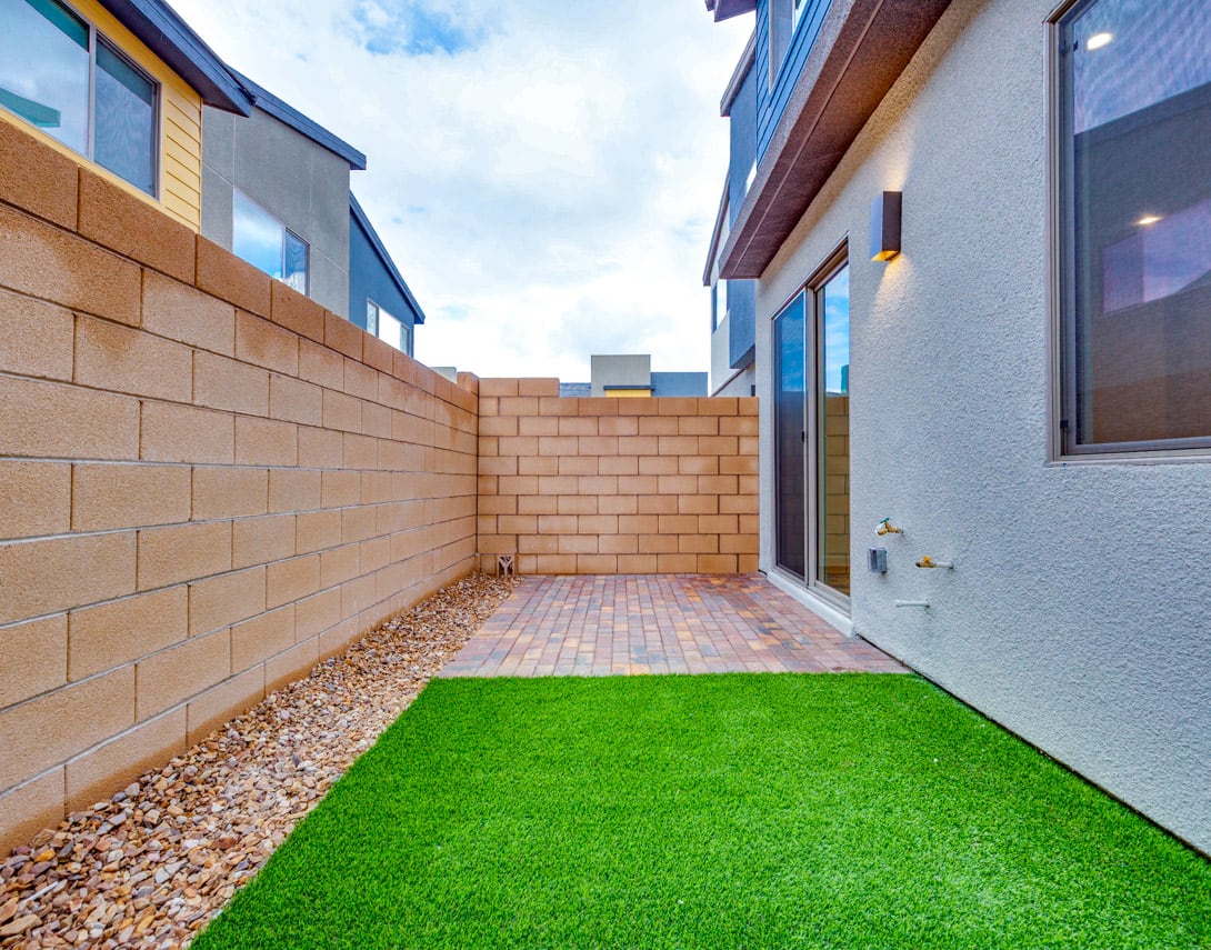A small, enclosed outdoor space with a brick wall, artificial grass, and gravel, surrounded by modern buildings with large windows.