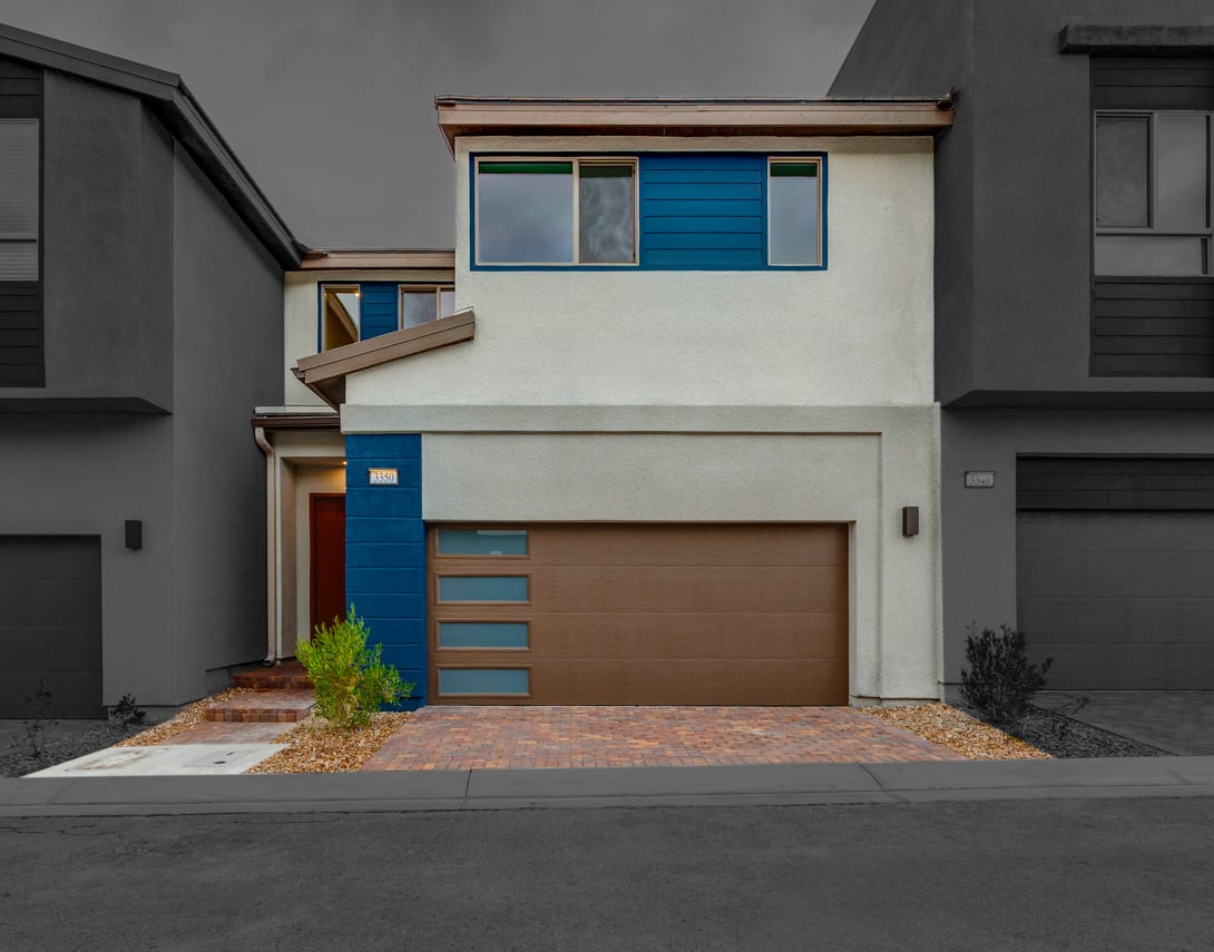 A modern, two-story residential building with a garage door, blue accents, and a brick walkway leading to the entrance.