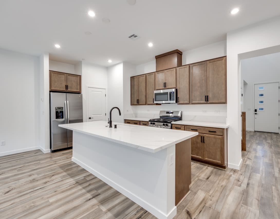 A modern, open-concept kitchen with white countertops, wooden cabinets, and stainless steel appliances, set against a backdrop of hardwood flooring.