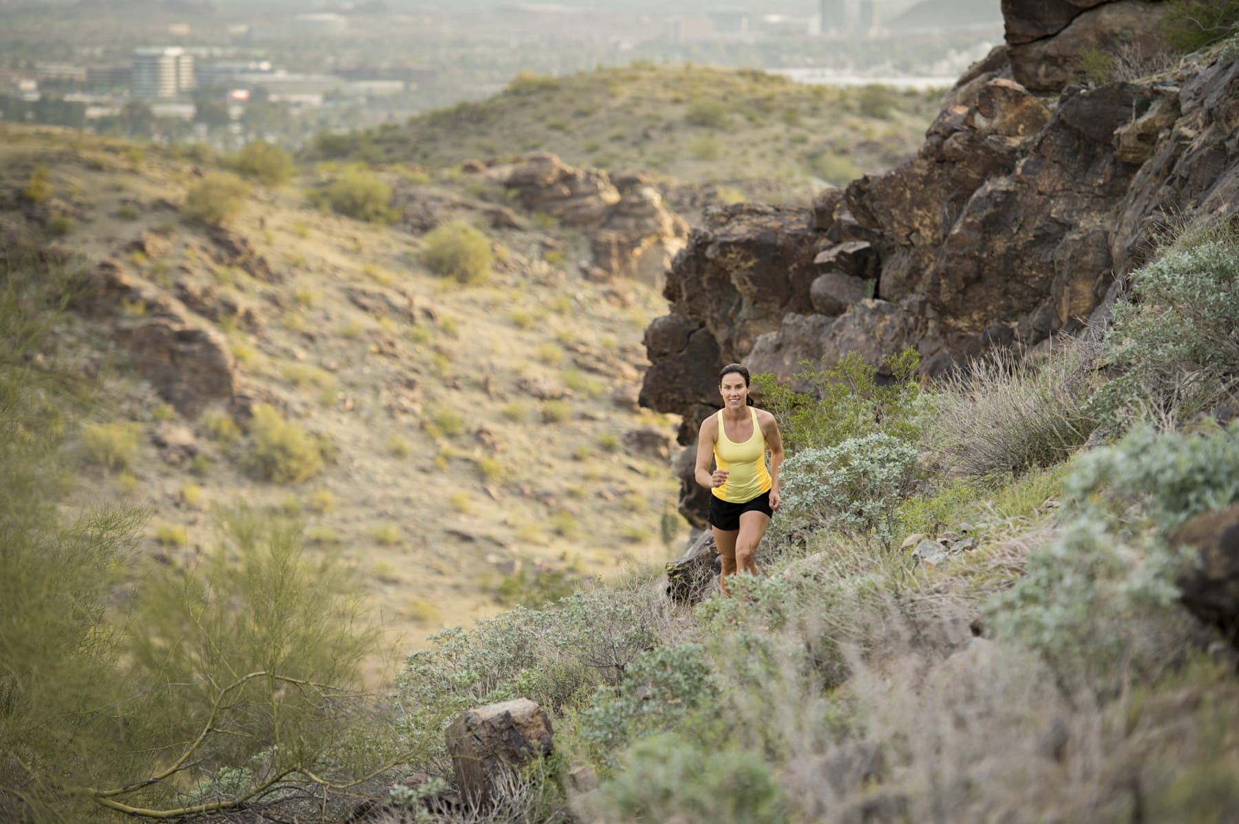 A person in athletic clothing is running on a rocky trail in a mountainous landscape with a city skyline visible in the distance.