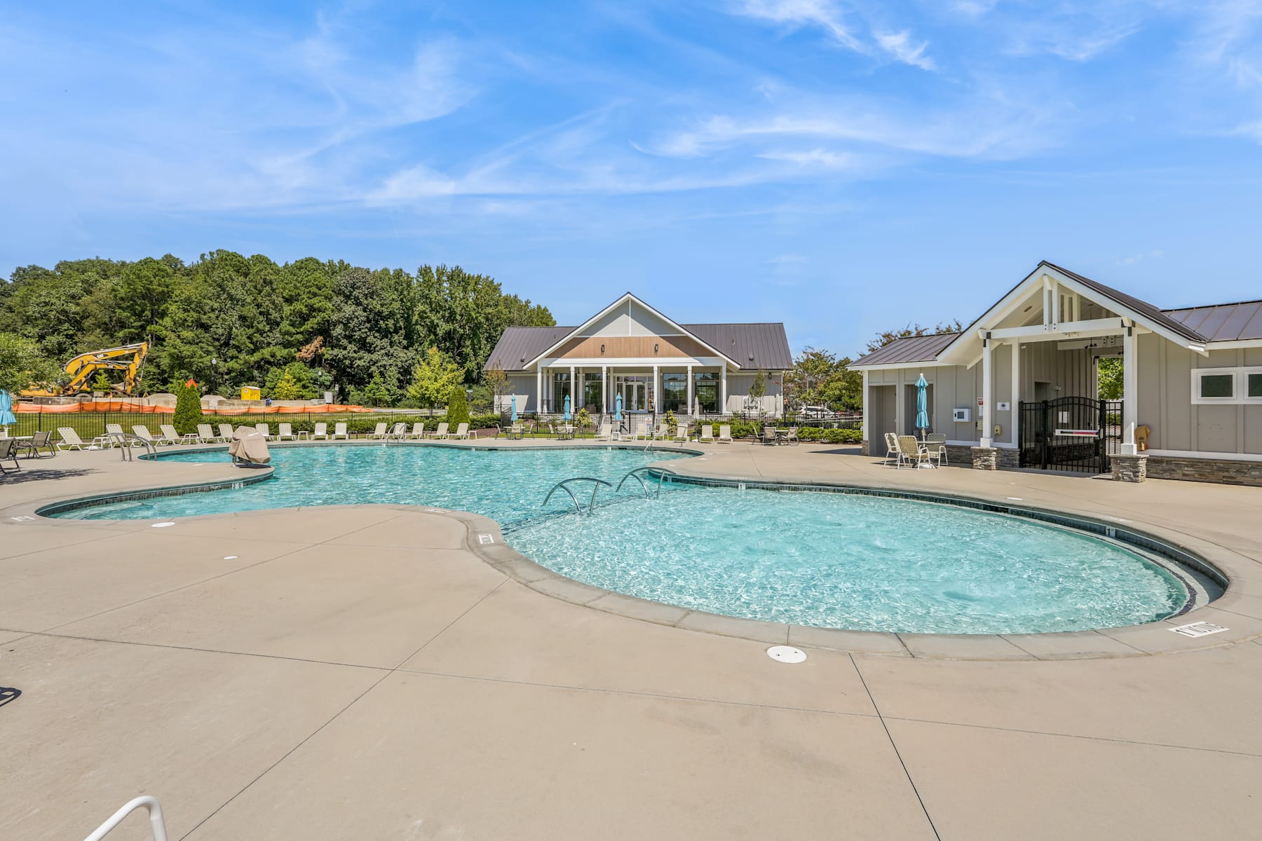 A large, inviting swimming pool surrounded by a paved deck, with a clubhouse-style building and lush greenery in the background.