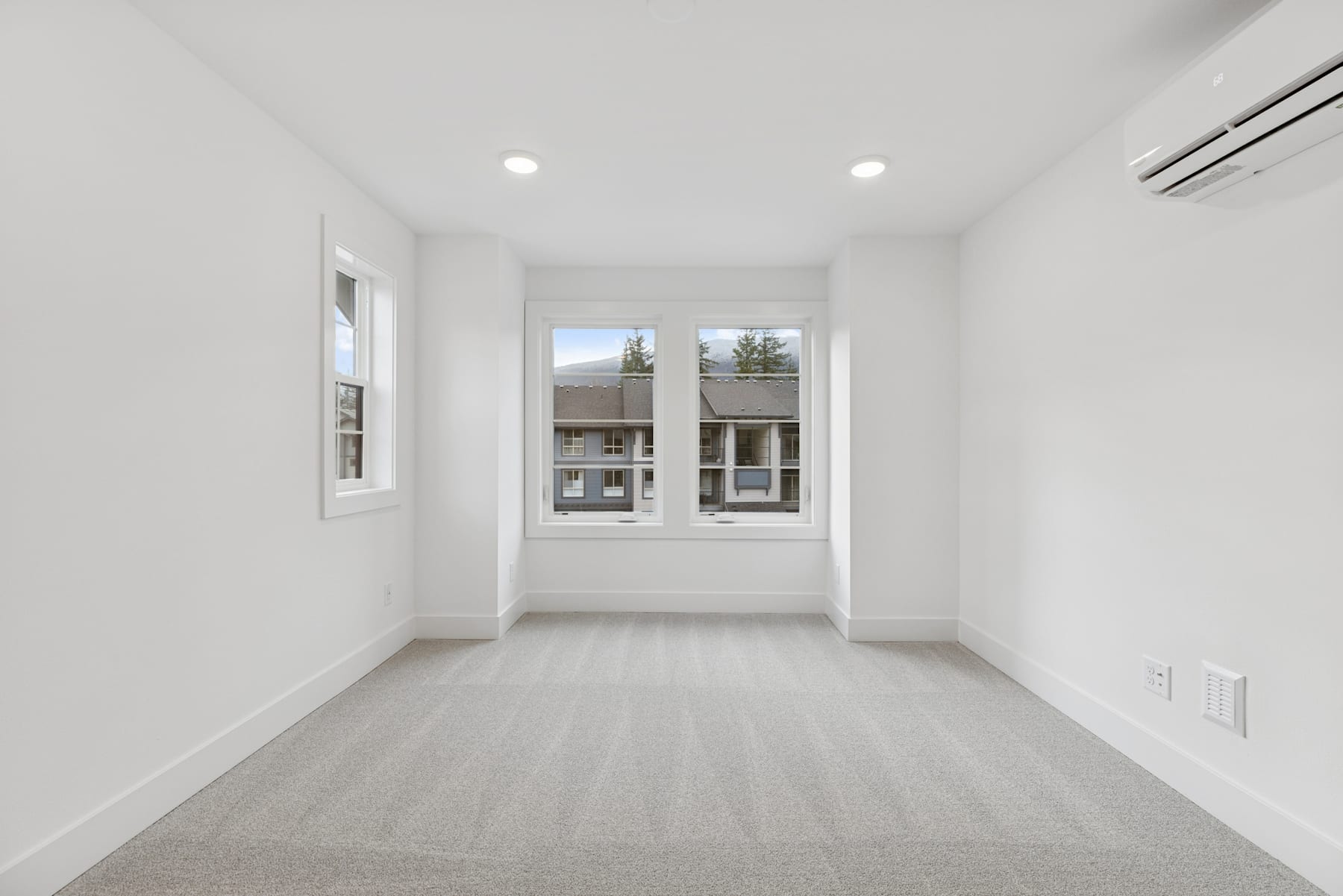 A bright, minimalist interior space with white walls, a hardwood floor, and large windows overlooking a wooded outdoor scene.