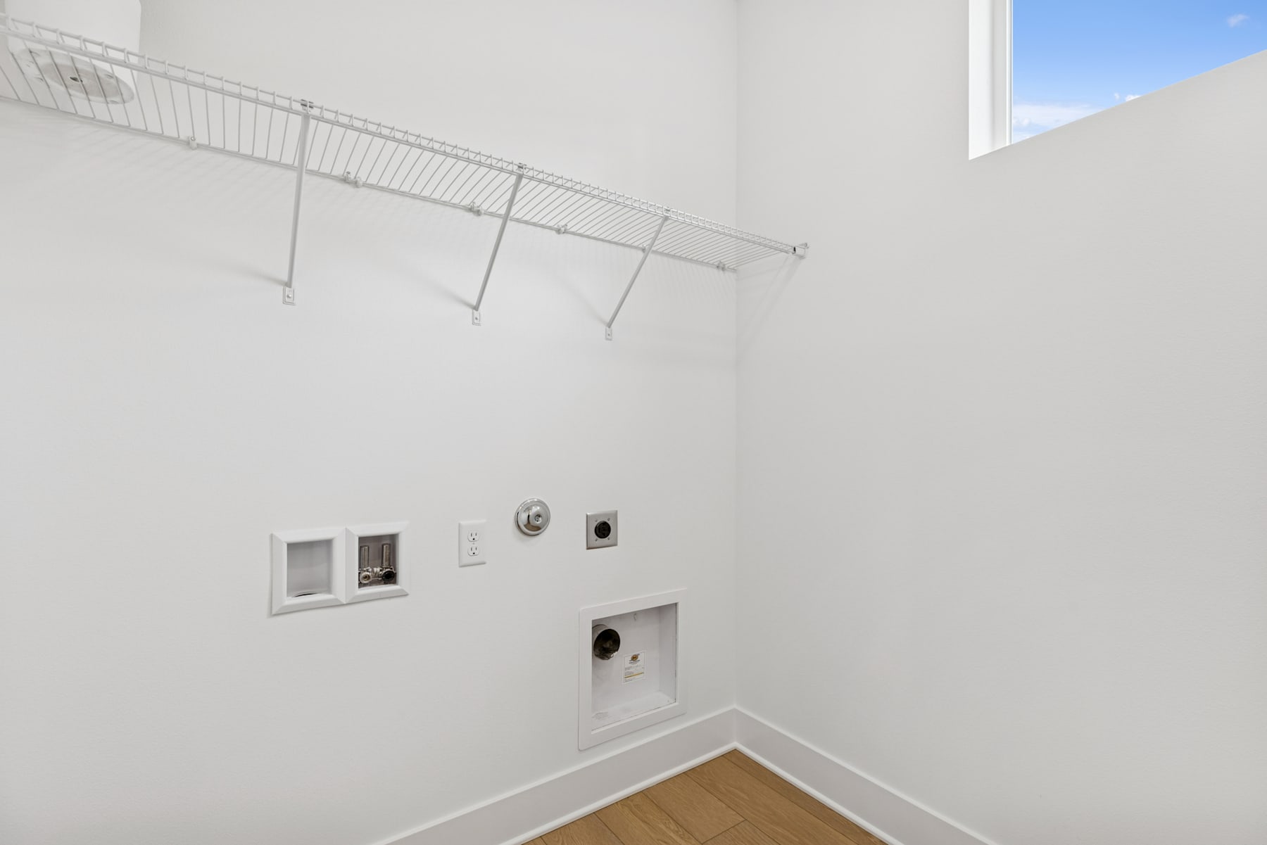 A minimalist, white-walled room with a metal clothing rack and electrical outlets visible in the foreground, and a window providing natural light in the background.
