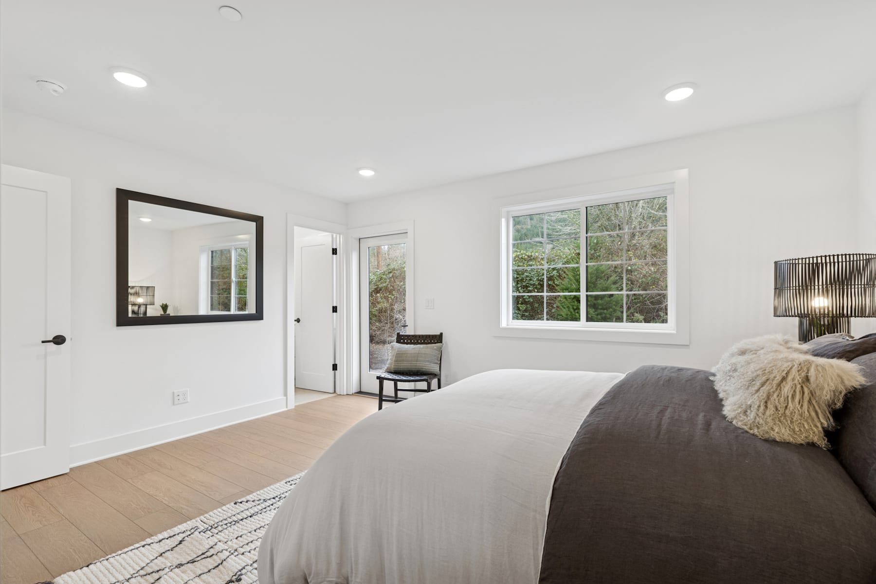 A cozy and well-lit bedroom with a large window, a plush gray comforter, and a black framed mirror on the wall.