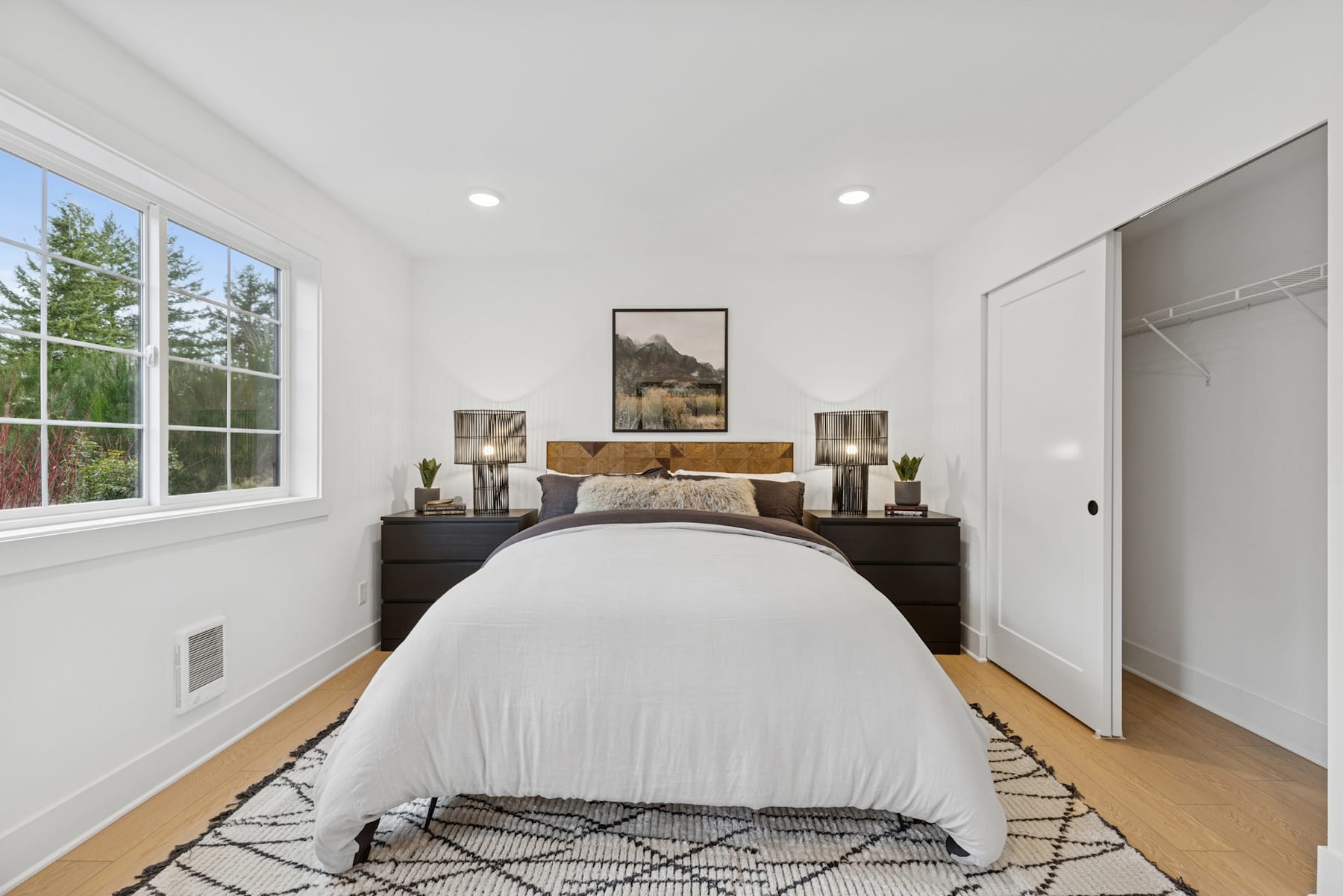 A cozy and well-designed bedroom with a large window overlooking a wooded landscape, featuring a plush bed, wooden furniture, and decorative accents.