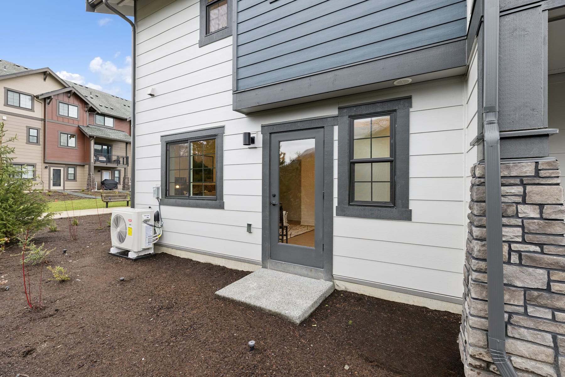 The image shows a modern, multi-story residential building with a stone and siding exterior, a covered entryway, and a landscaped yard in the foreground.