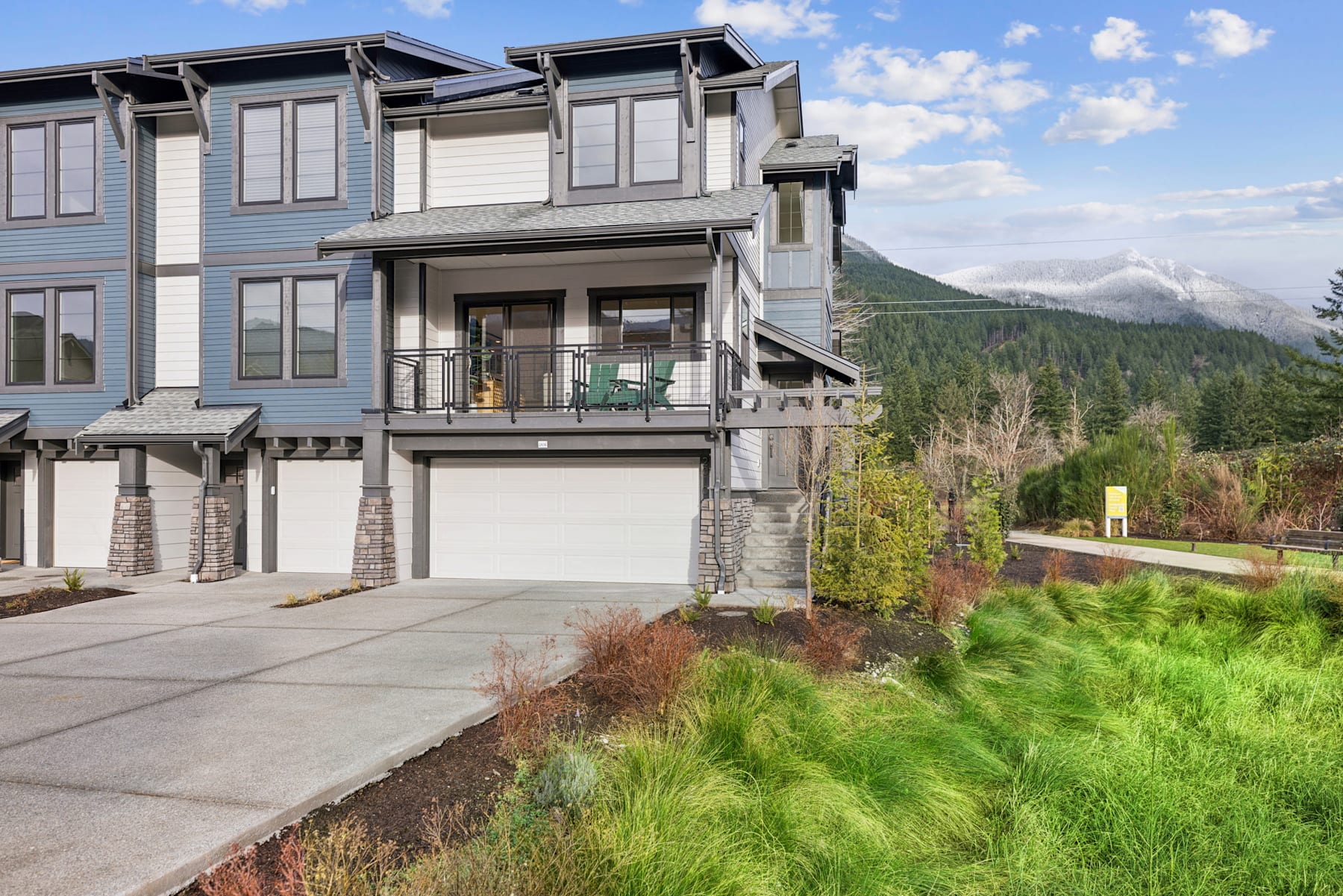 A modern multi-story townhouse with a balcony and garage, situated against a backdrop of lush greenery and mountains in the distance.