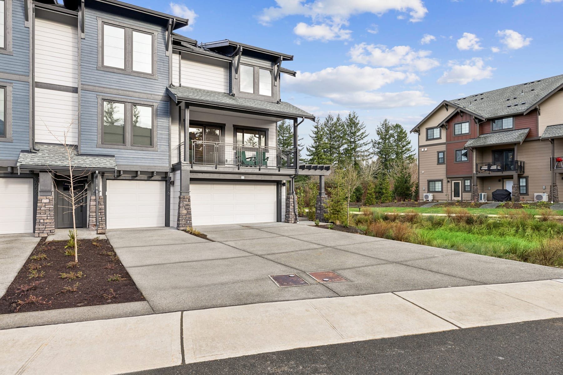 The image depicts a residential neighborhood with modern townhouses, a paved driveway, and a grassy area in the foreground. The buildings have a mix of siding materials and architectural styles, and the sky is filled with fluffy white clouds.