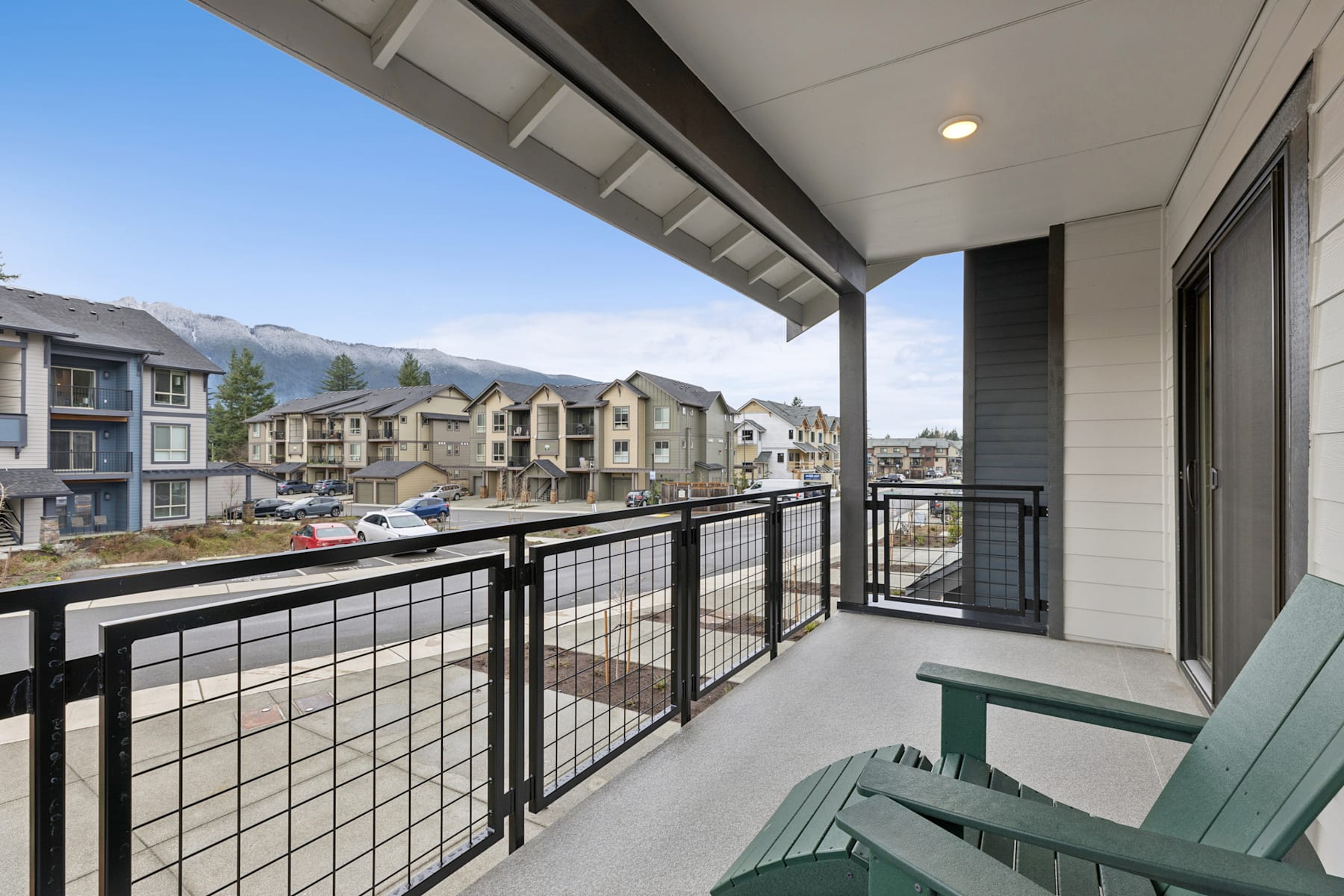 A balcony with a metal railing overlooks a residential neighborhood with multi-story apartment buildings in the background, set against a clear blue sky.