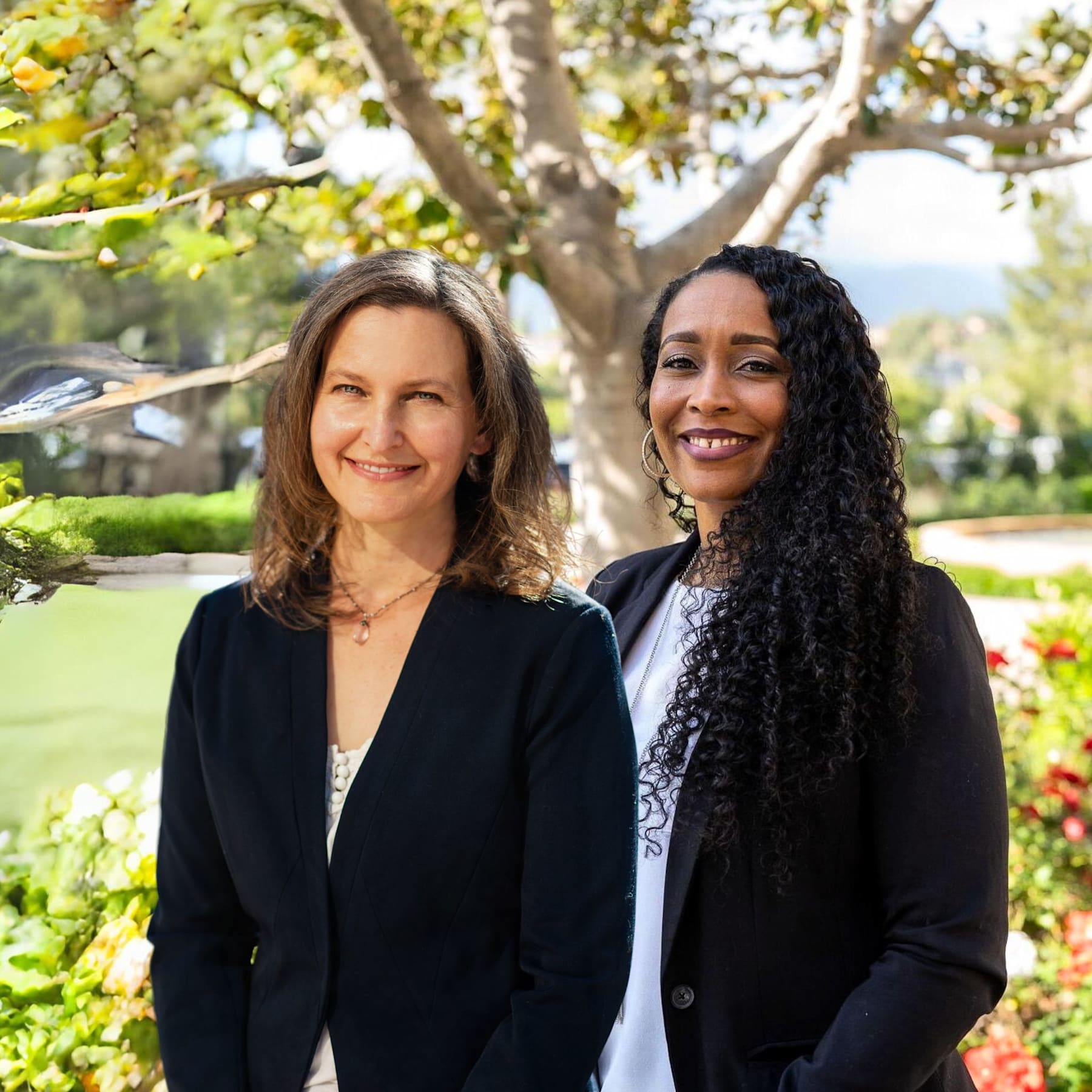 Two women, one with brown hair and the other with curly dark hair, are standing together in a lush, green outdoor setting with trees and flowers in the background.