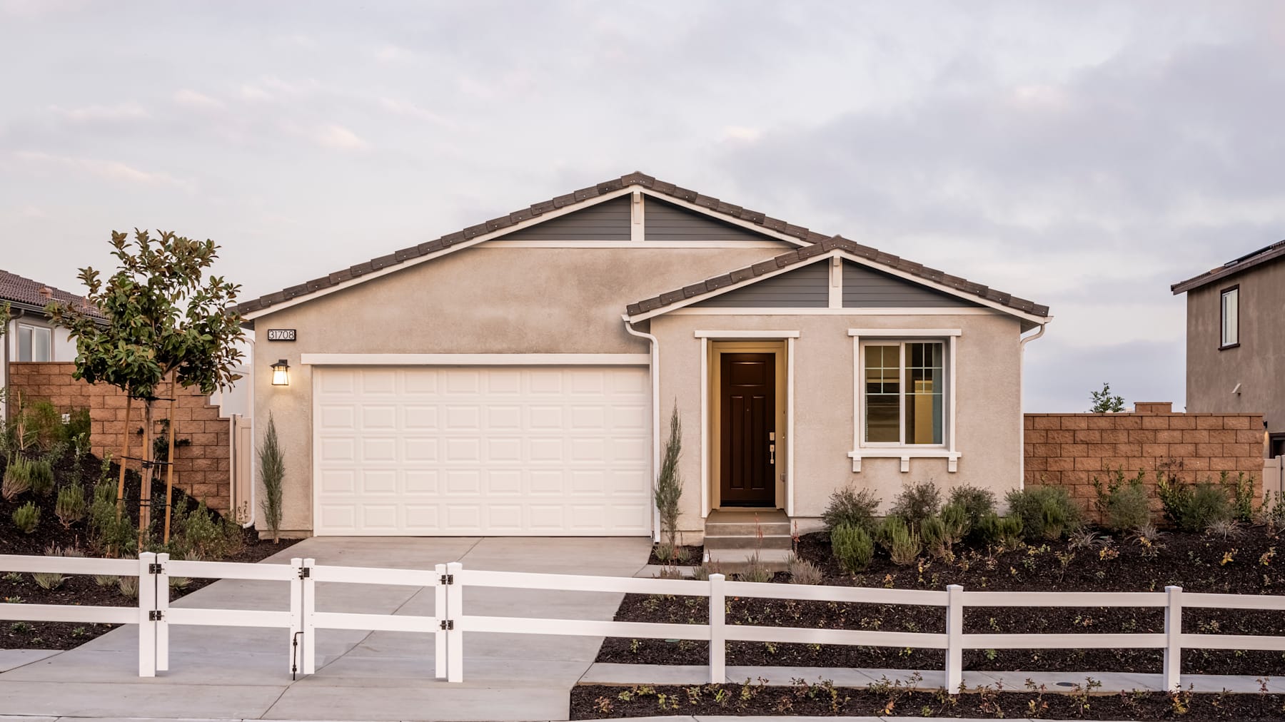 A single-story beige house with a garage door and a white picket fence in the foreground, set against a cloudy sky in the background.