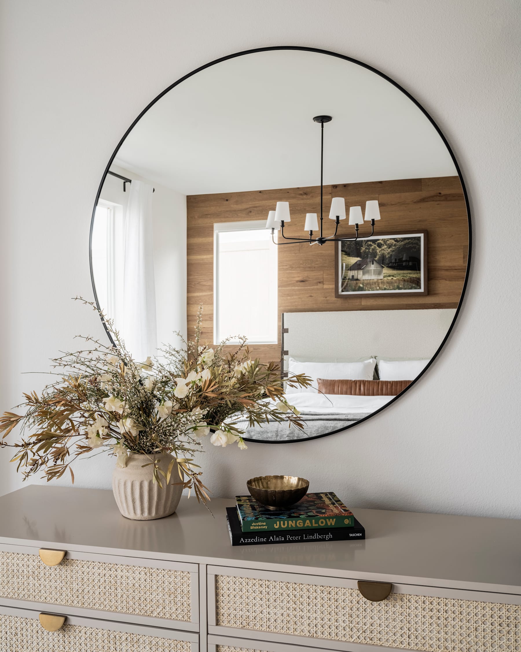 A large, oval-shaped mirror hangs on the wall, reflecting a cozy living room with a wooden accent wall, a chandelier, and a decorative floral arrangement on a dresser in the foreground.