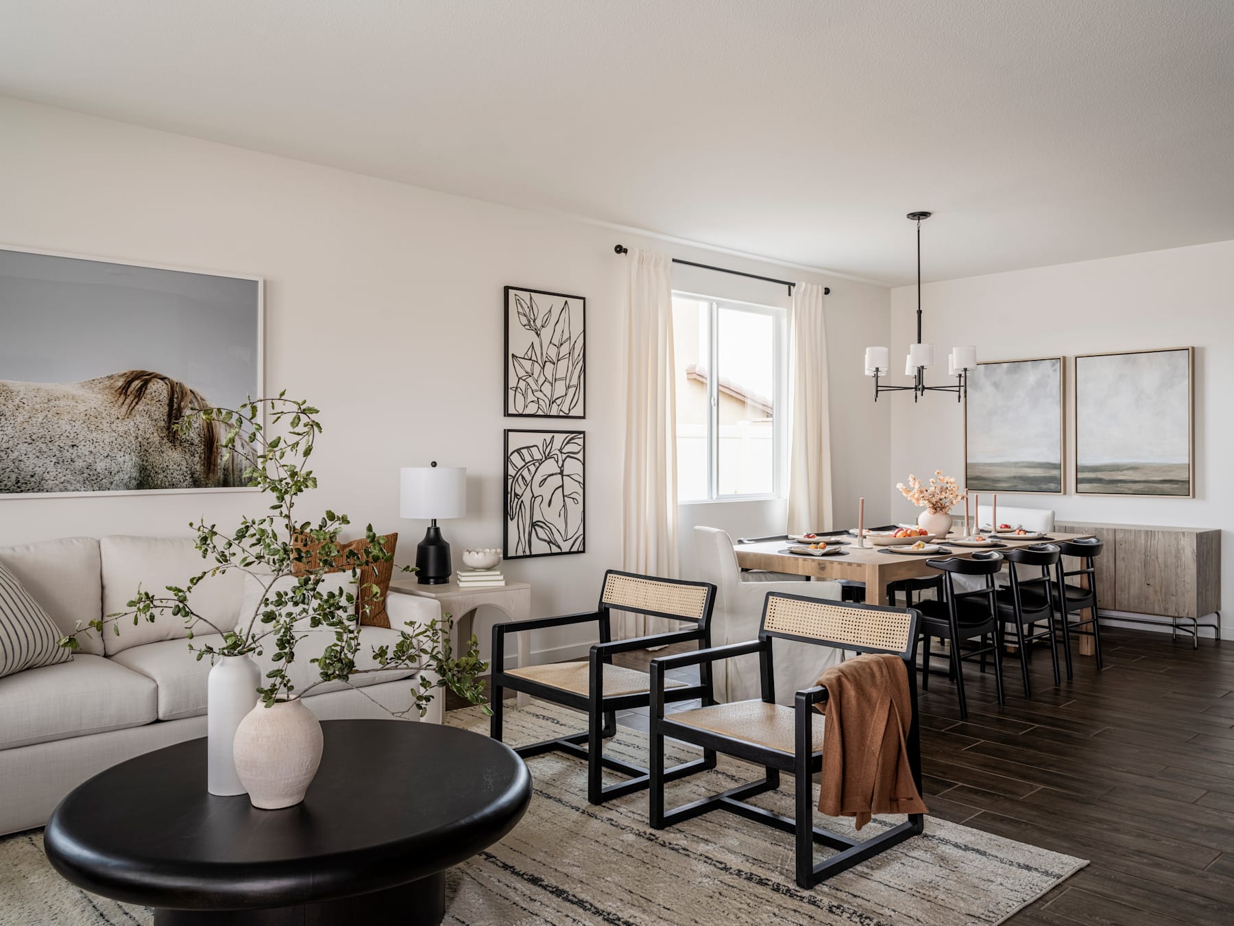 A cozy and stylish living room with a dining area, featuring a gray sofa, a round black coffee table, and various decorative elements such as wall art and potted plants.