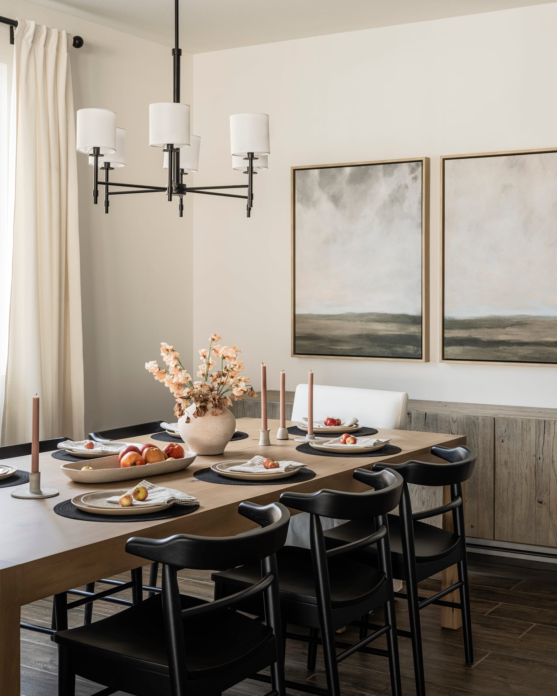 A cozy and inviting dining room with a wooden table set for a meal, surrounded by black chairs and adorned with a floral arrangement, framed artwork, and a modern chandelier.
