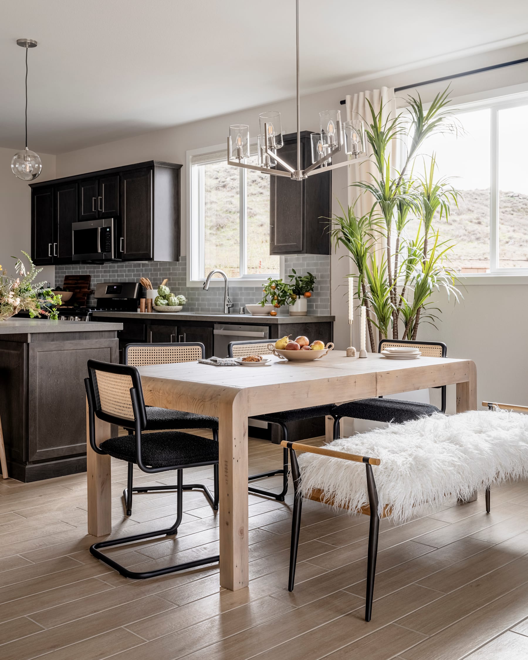 A modern and stylish kitchen with dark cabinets, a wooden dining table, and a cozy seating area with a furry bench, all set against a backdrop of large windows and lush greenery.