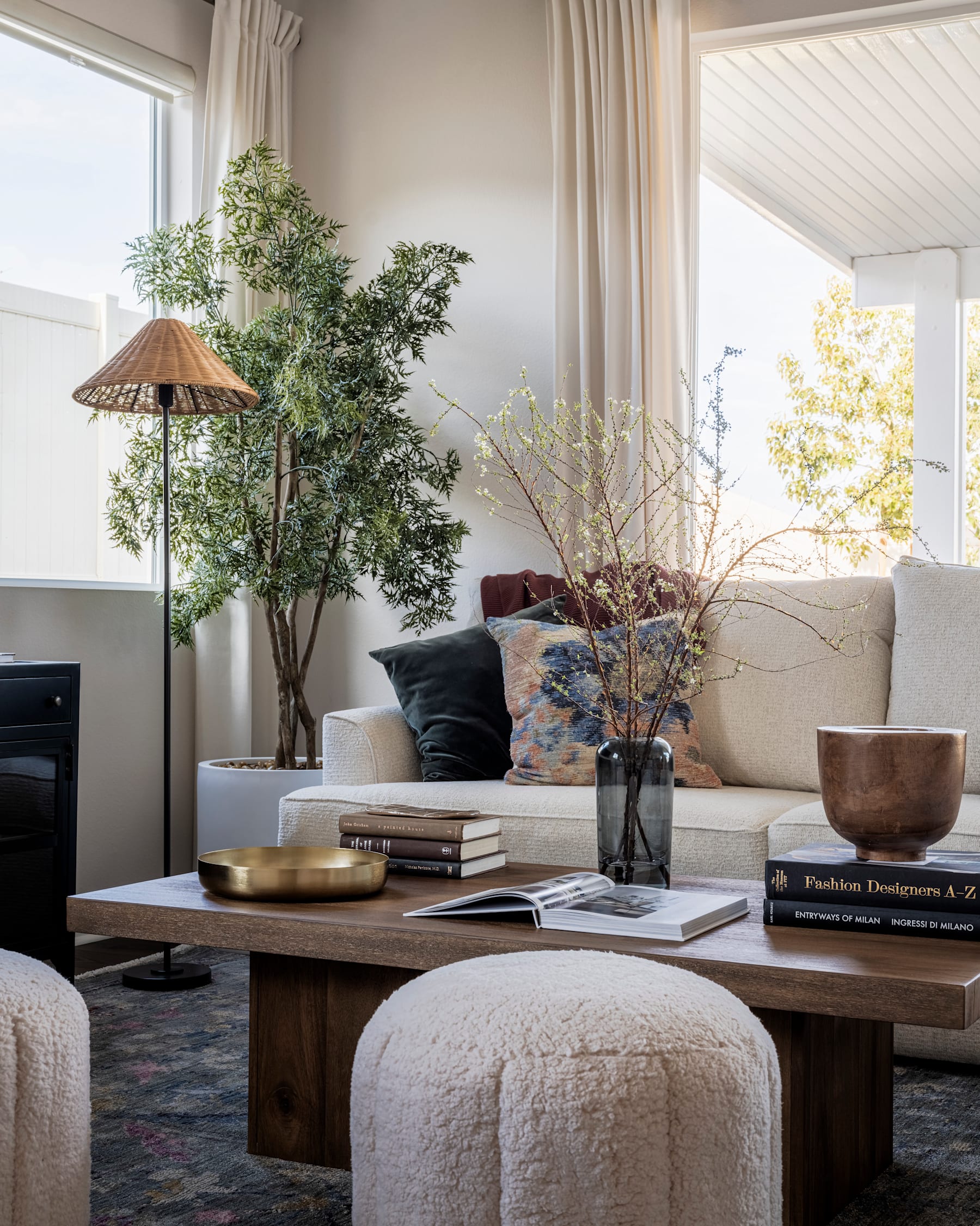 A cozy and inviting living room with a large wooden coffee table, a potted plant, and a vase of flowers, set against a backdrop of large windows and curtains.