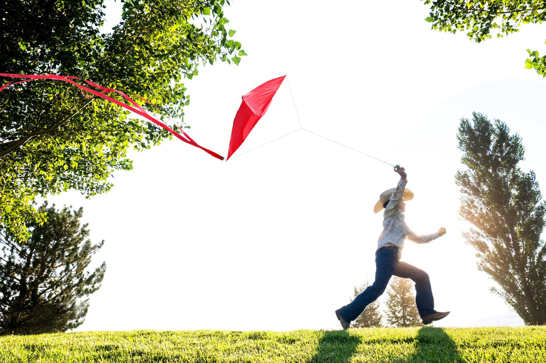 A person is running on a grassy field, with a red kite flying above them amidst the lush greenery and trees in the background.
