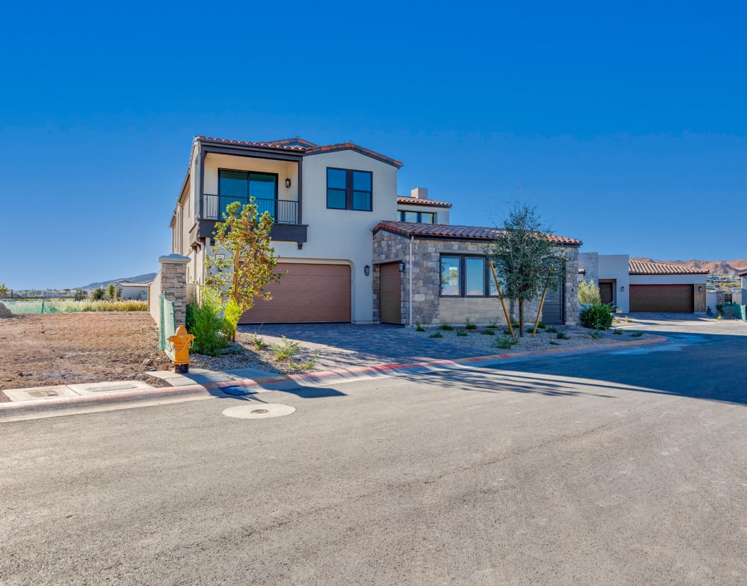 A modern, two-story residential house with a tile roof, surrounded by a paved driveway and landscaped yard against a clear blue sky.
