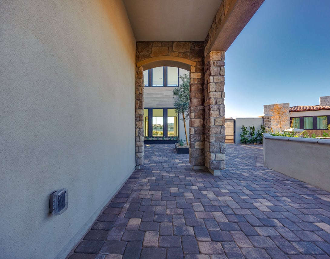 A paved walkway leads through an archway in a stone wall, with a building visible in the background and a clear blue sky overhead.