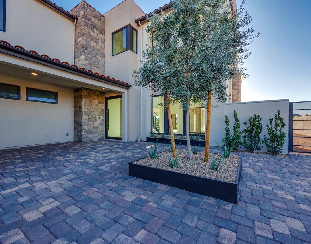 A modern, two-story house with a paved courtyard in the foreground, featuring a large olive tree and decorative planters. The building has a mix of stone and stucco exterior, with large windows and a tiled roof.