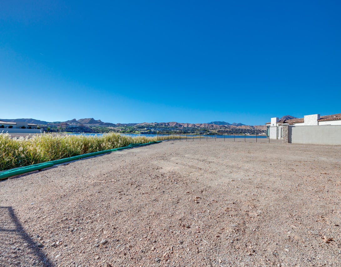A gravel path leads to a scenic view of a body of water surrounded by mountains and buildings in the background under a clear blue sky.