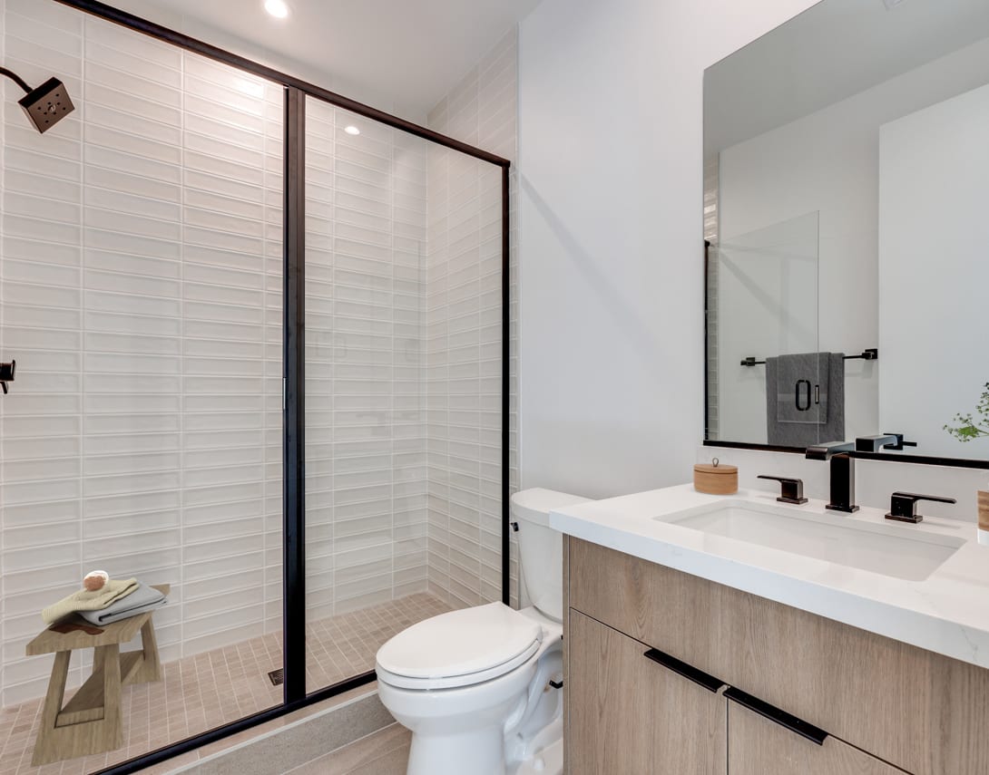 A modern bathroom with a white tiled wall, a wooden vanity with a white countertop, and a black-framed glass shower enclosure.