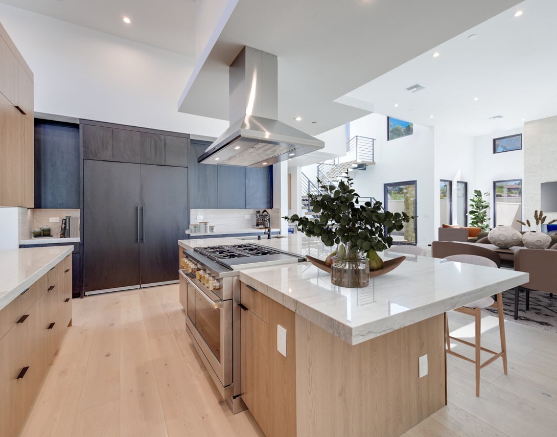 A modern, open-concept kitchen with dark cabinets, a large island with a white countertop, and a range hood above the stove. The kitchen is part of a larger living space with natural light streaming in through the windows.