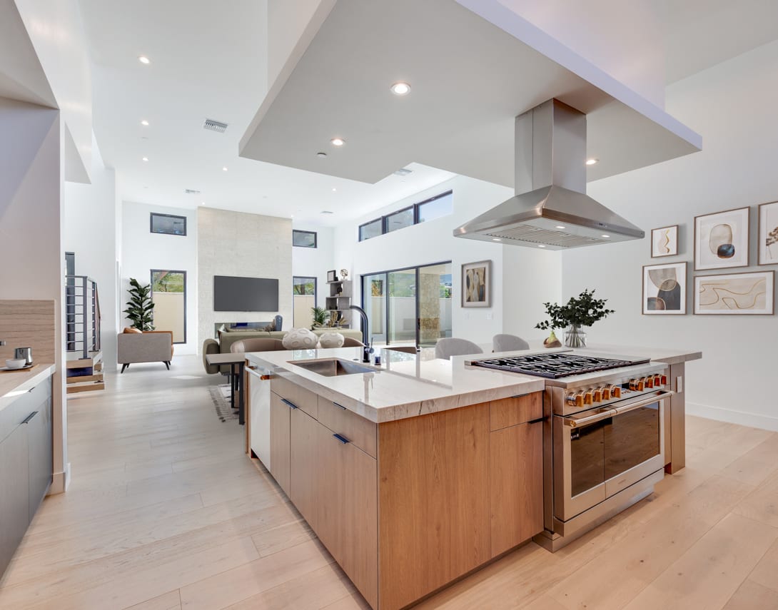 A modern, open-concept kitchen and living room with a large island, stainless steel appliances, and natural wood cabinetry, set against a bright, airy backdrop with high ceilings and large windows.