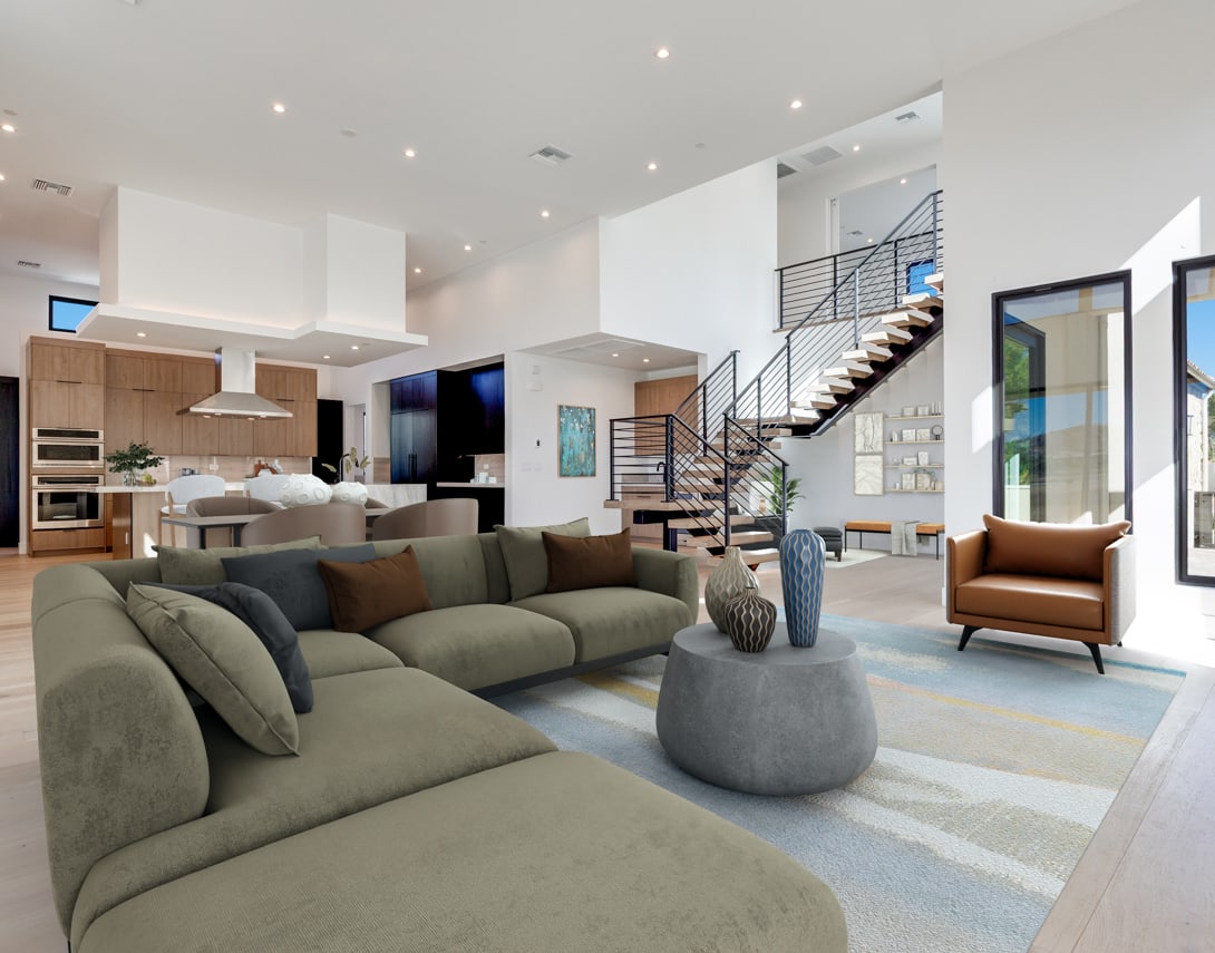 A spacious and modern living room with a large green sofa, a gray circular ottoman, and a staircase leading to an upper level. The room features a kitchen area with wooden cabinets and stainless steel appliances, and large windows providing natural light.