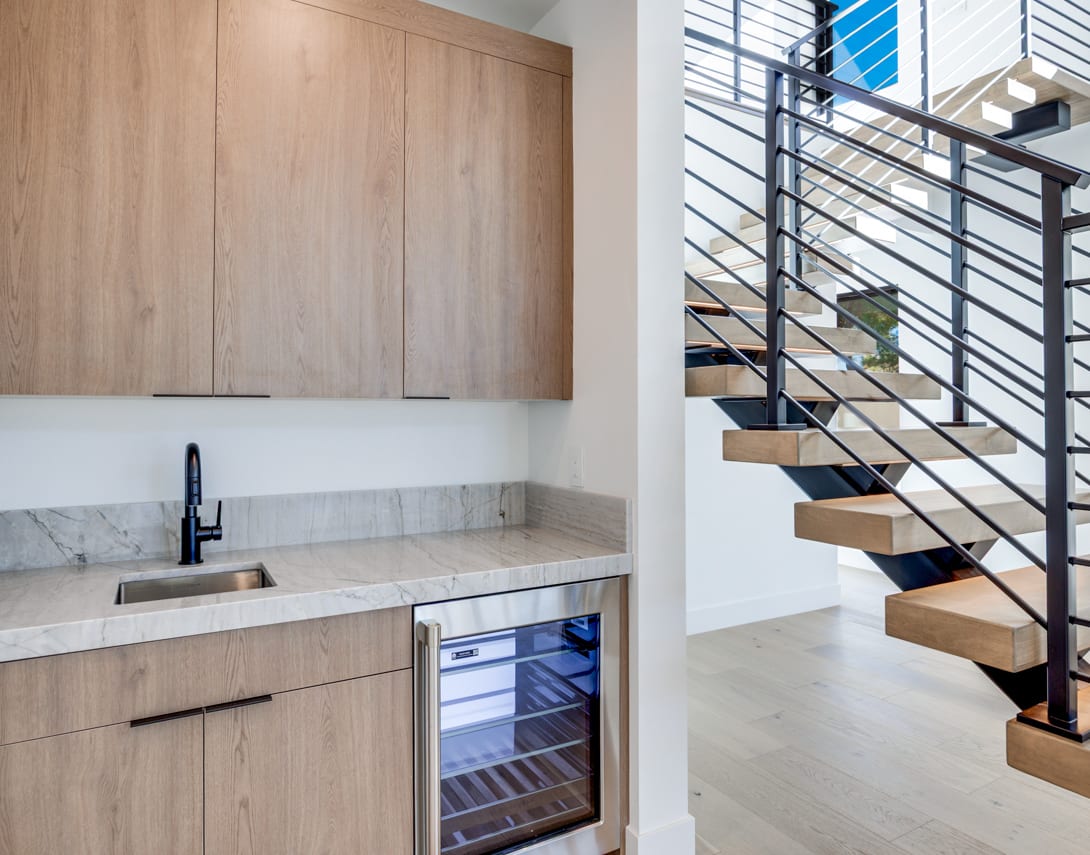 A modern kitchen with wooden cabinets and a marble countertop, adjacent to a staircase with metal railings leading to an upper level.