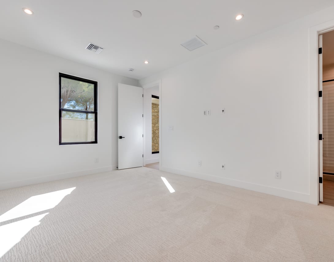 A bright, spacious room with white walls, a large window, and a carpeted floor. The room appears to be empty, with a framed artwork hanging on the wall and a door visible in the background.