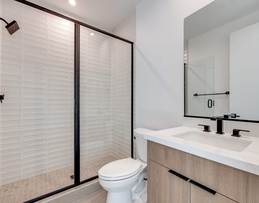 A modern bathroom with a white vanity, a toilet, and a glass-enclosed shower area with white tiled walls.