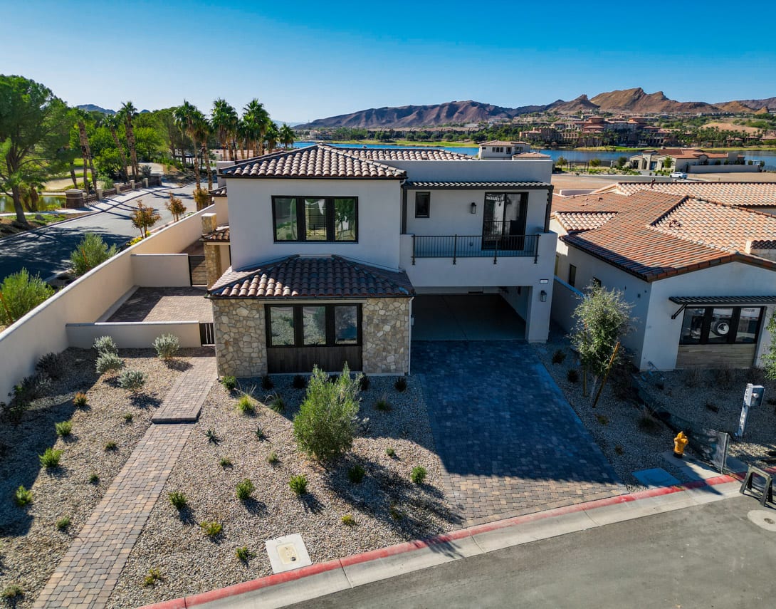 A modern, multi-story residential building with a tiled roof and stone exterior sits amidst a landscaped courtyard, surrounded by lush greenery and mountains in the background.