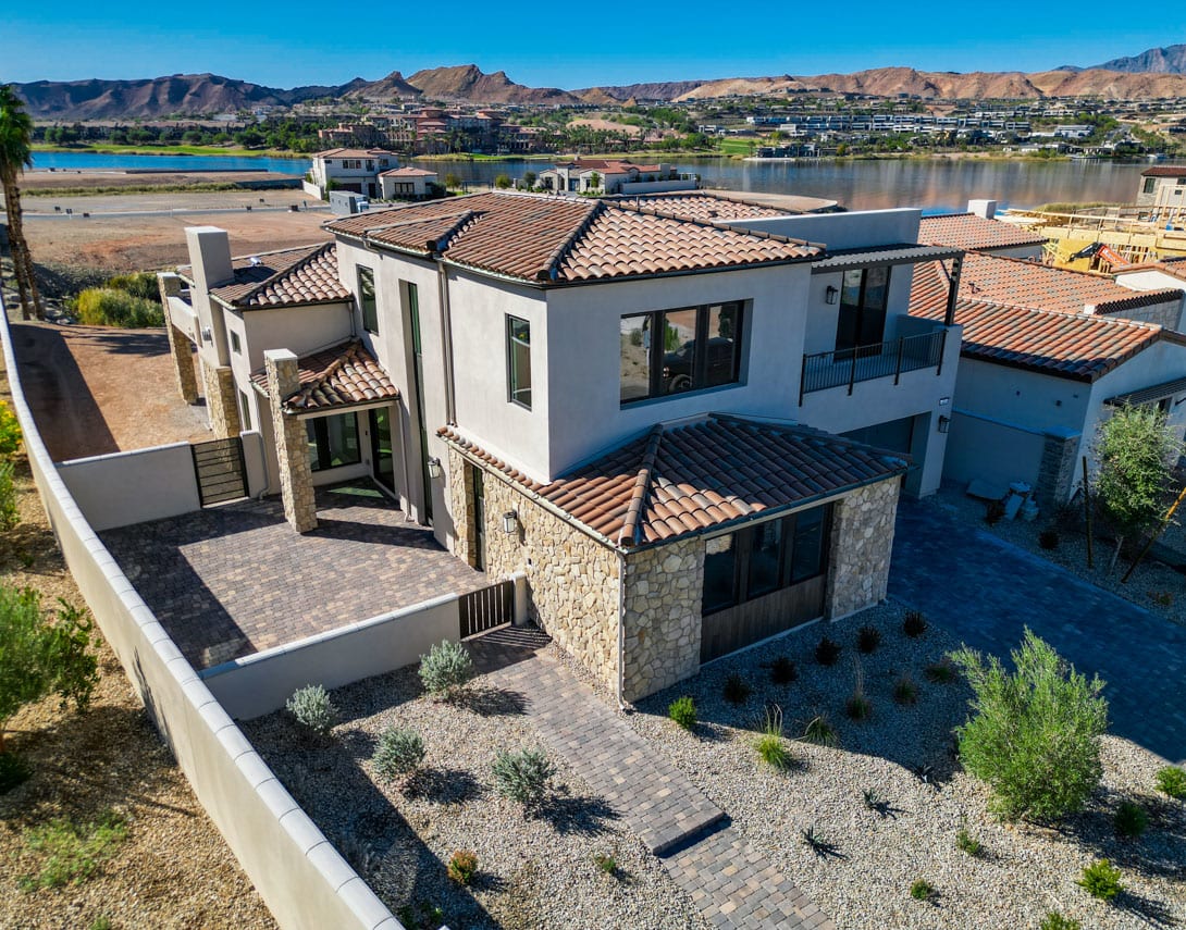 A large, multi-story Mediterranean-style house with a tiled roof sits atop a hill, overlooking a scenic desert landscape with mountains in the background.