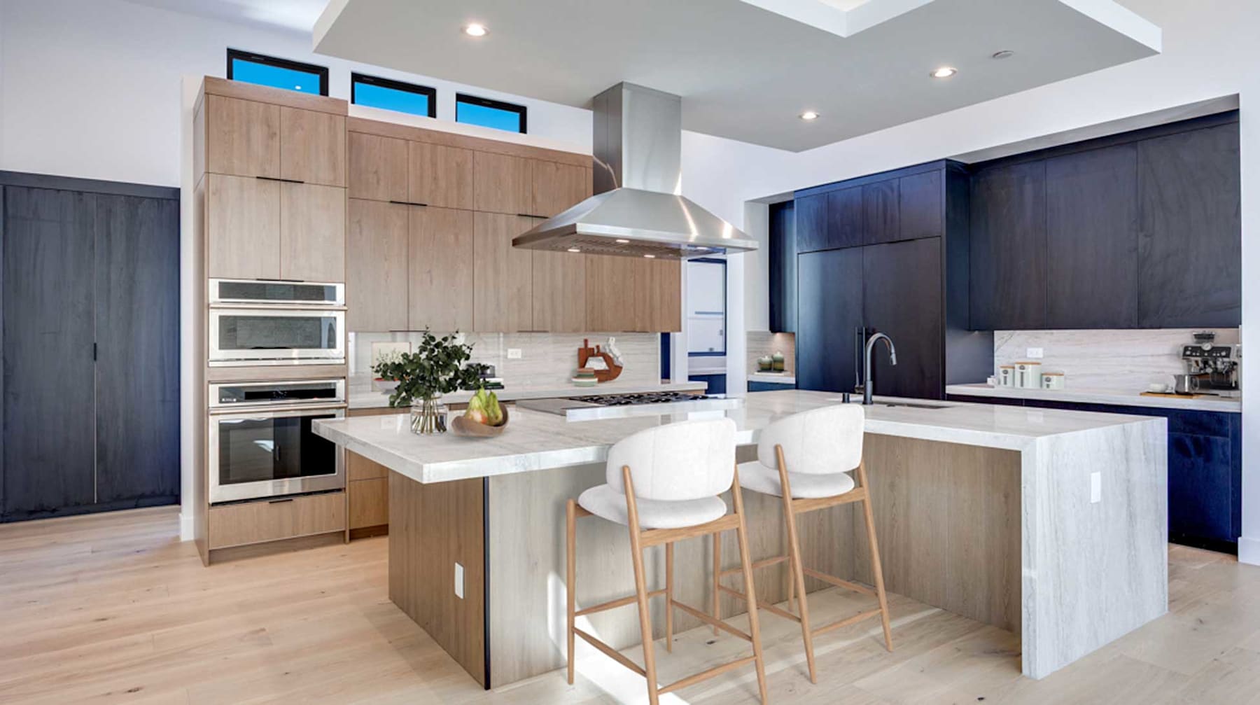 A modern and sleek kitchen with light wood cabinets, a white countertop, and bar stools, set against a backdrop of dark cabinetry and stainless steel appliances.