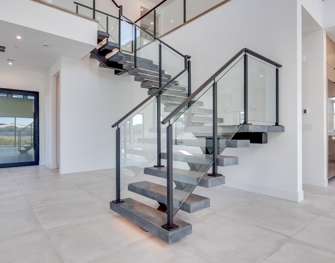 A modern, minimalist staircase with glass panels and metal railings stands in a bright, open-concept interior with white walls and a tiled floor.