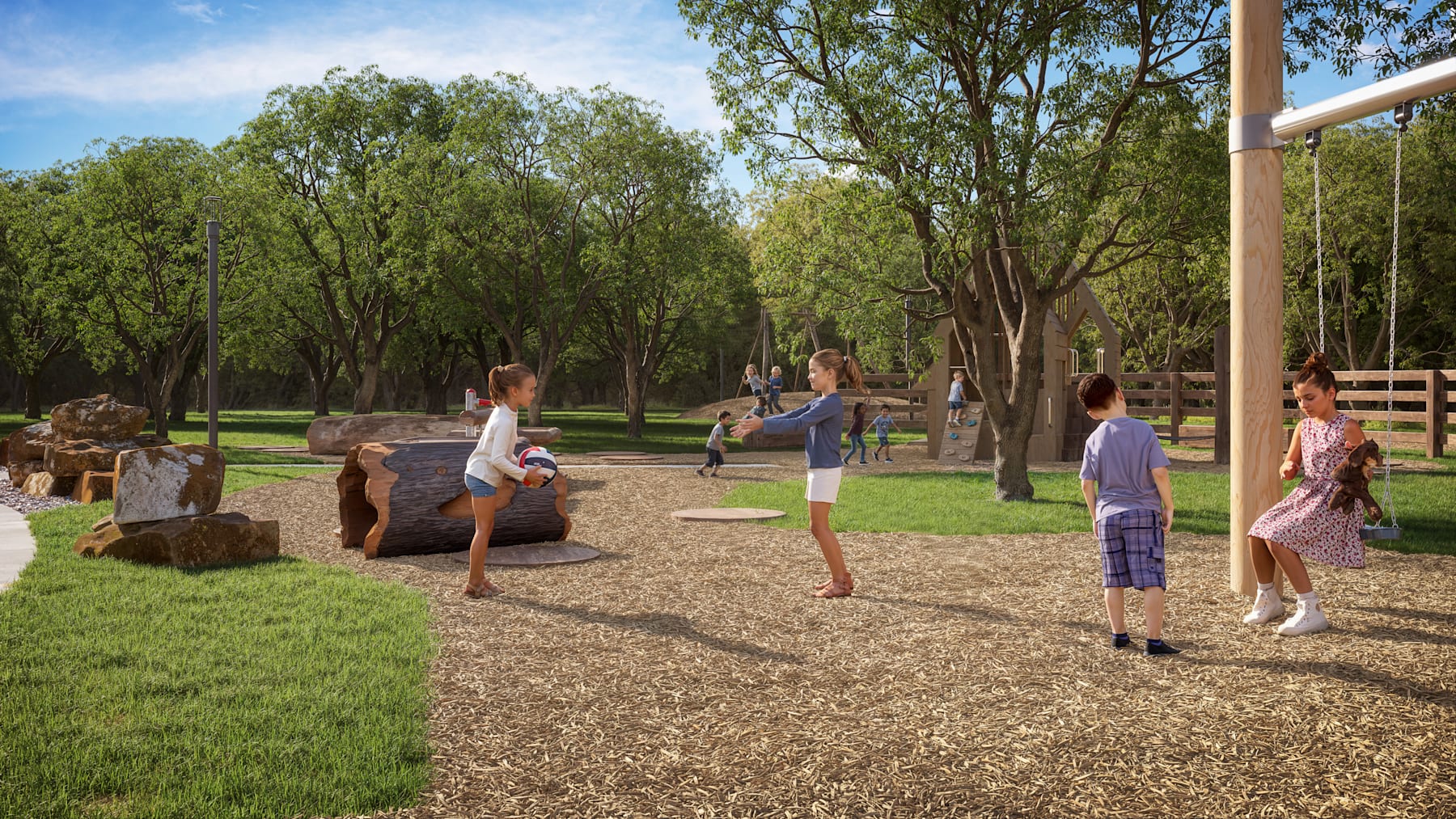 A group of people, both adults and children, are gathered in a park-like setting with lush greenery, trees, and a grassy area with wood chips on the ground.