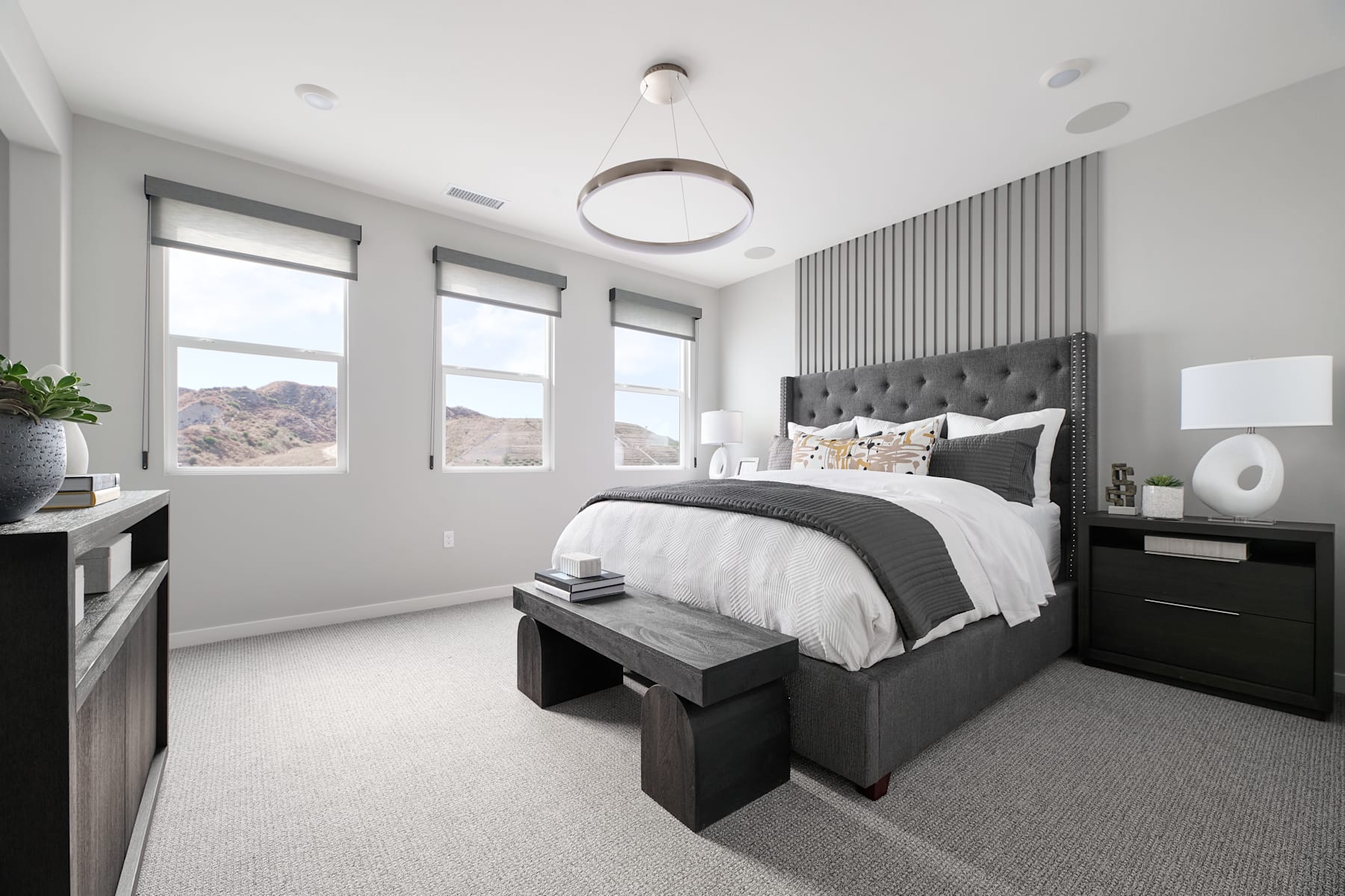 A modern and minimalist bedroom with a large bed, dark furniture, and large windows overlooking a scenic outdoor landscape.