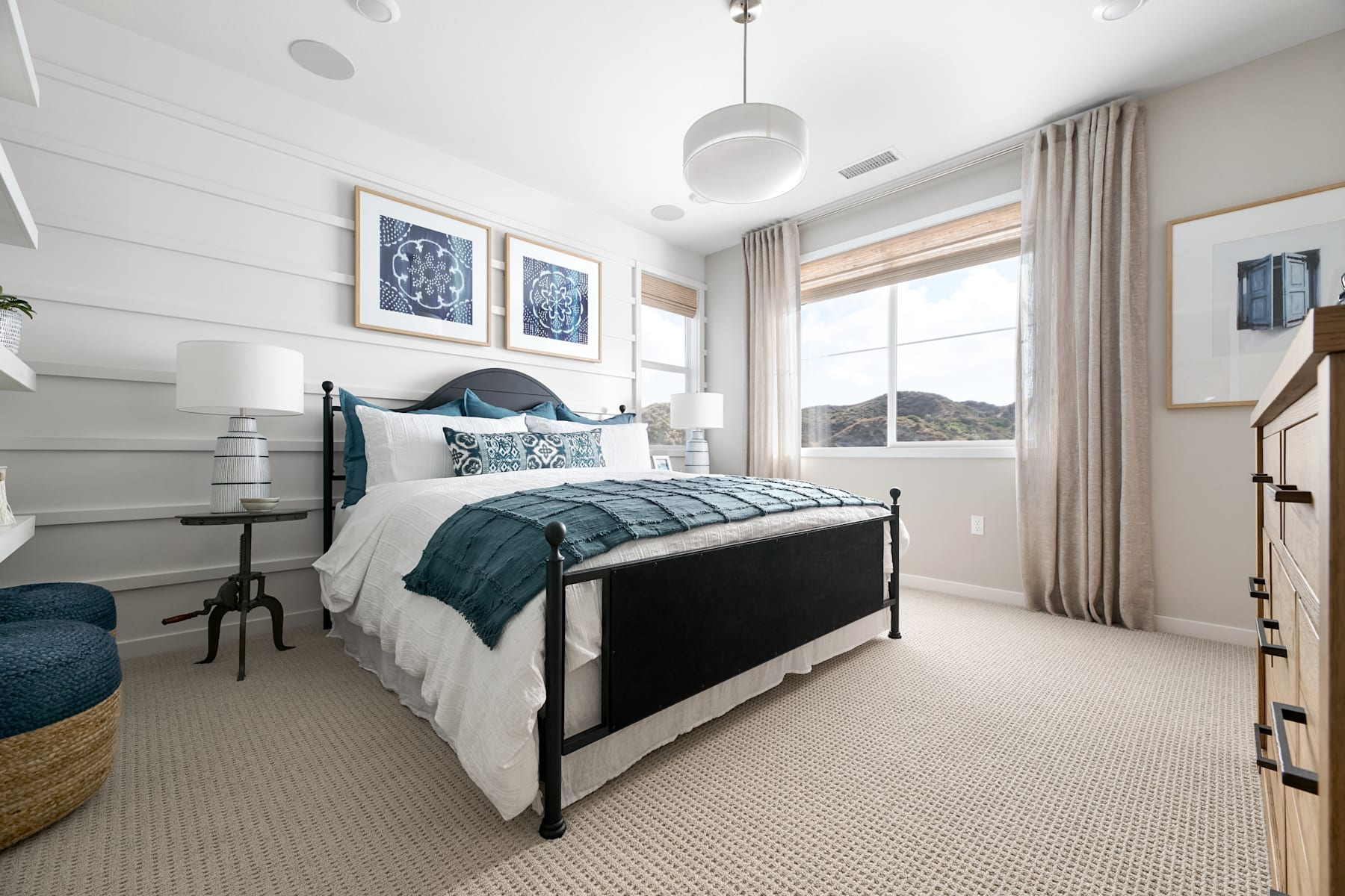 A cozy and well-decorated bedroom with a large bed, patterned bedding, and framed artwork on the walls, set against a backdrop of large windows overlooking a scenic outdoor landscape.