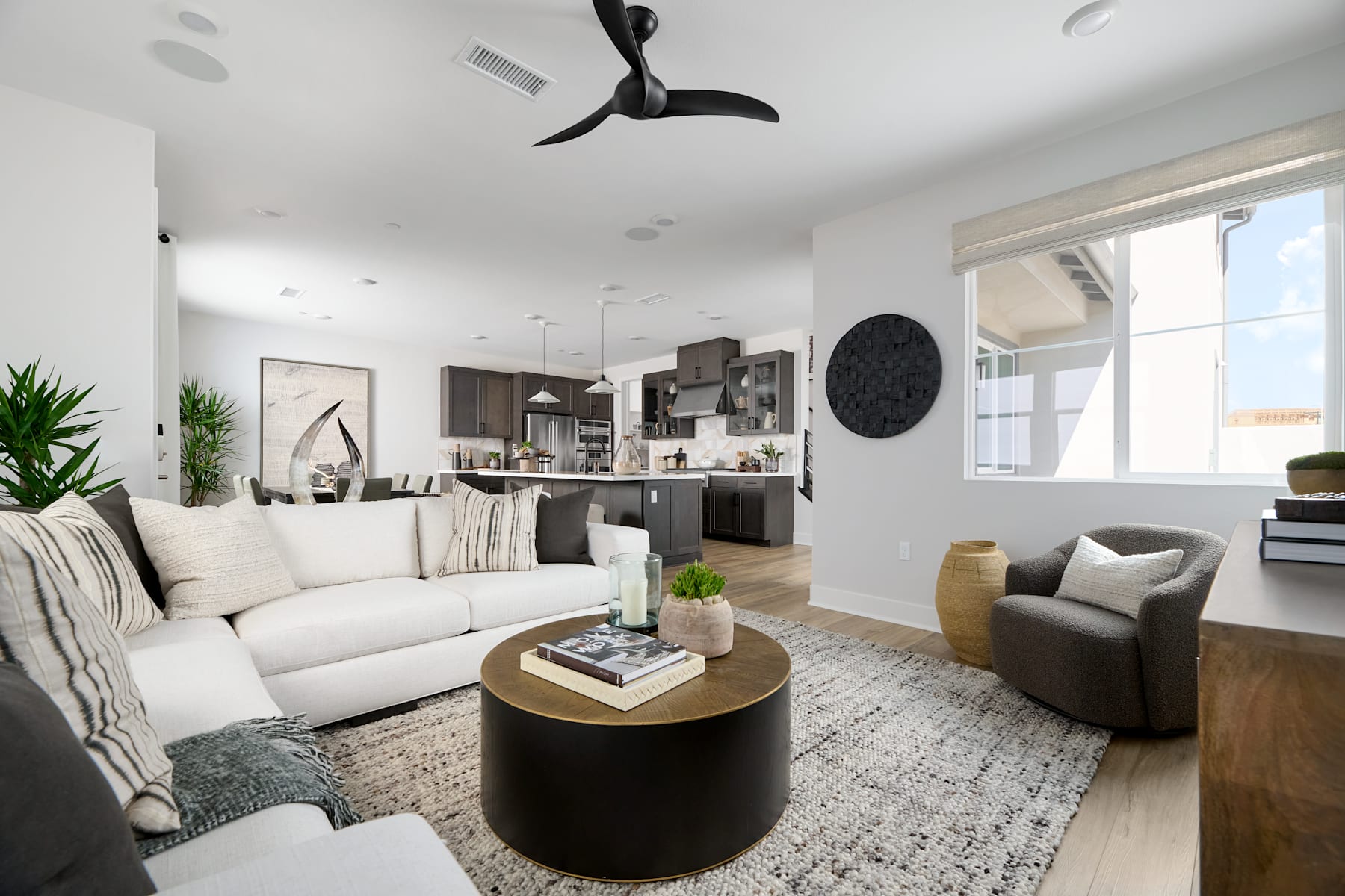 A cozy and modern living room with a white sofa, a round wooden coffee table, and a ceiling fan, surrounded by various decorative elements and a view of the kitchen area in the background.