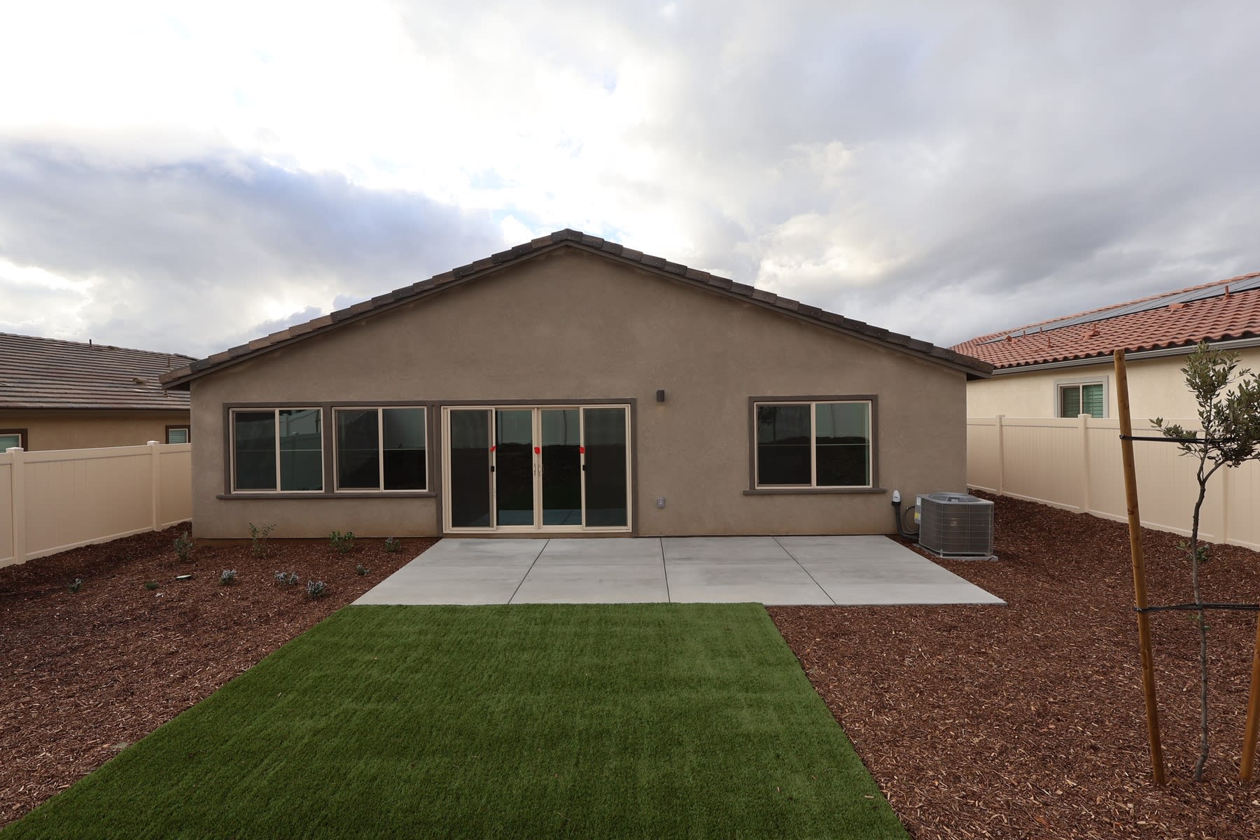 A single-story house with a tiled roof, surrounded by a concrete patio, a grassy lawn, and a gravel-covered area in the foreground.
