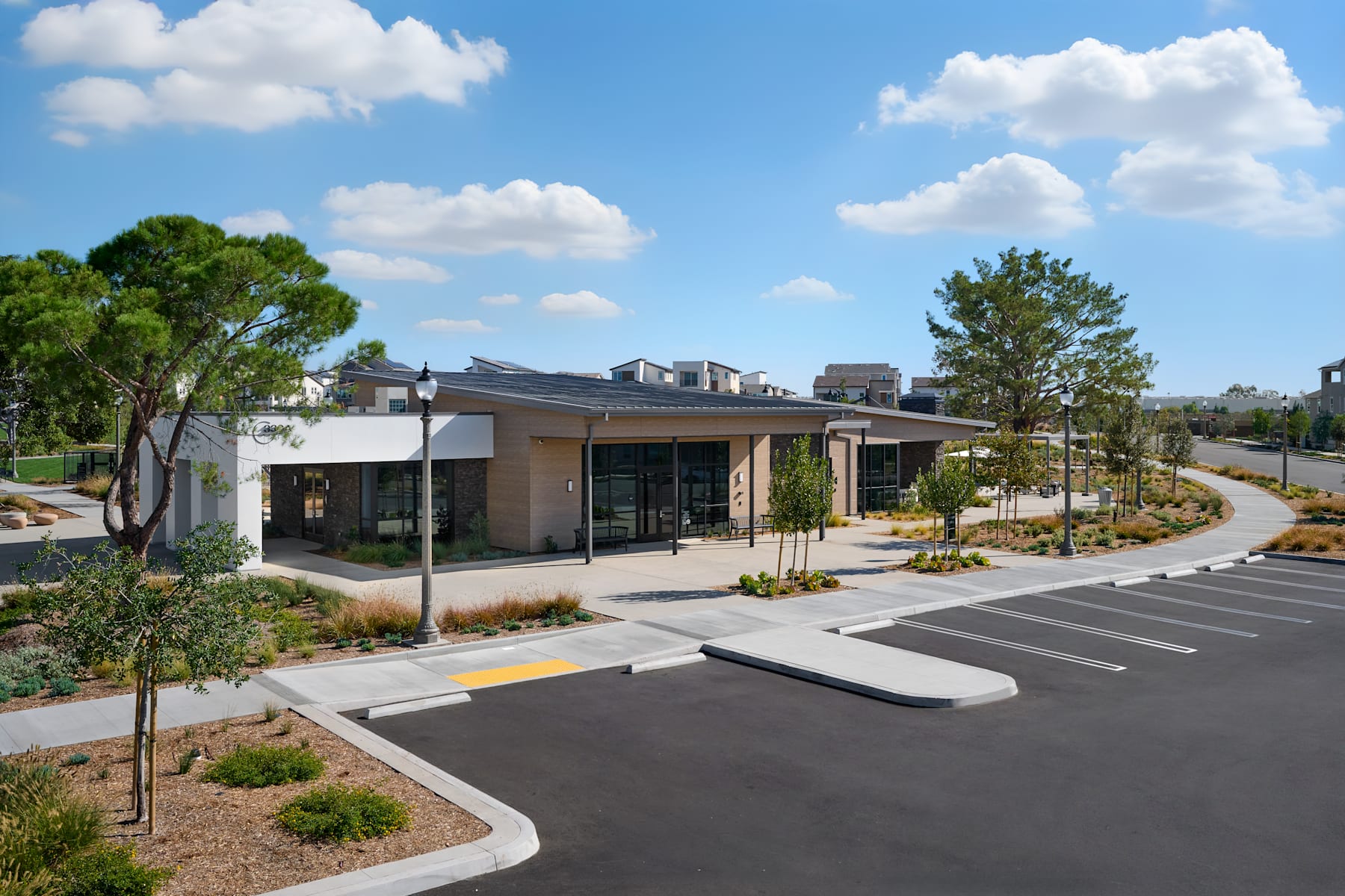 A modern, single-story building with large glass windows and a covered entrance sits in a landscaped area with trees, shrubs, and a paved parking lot in the foreground.