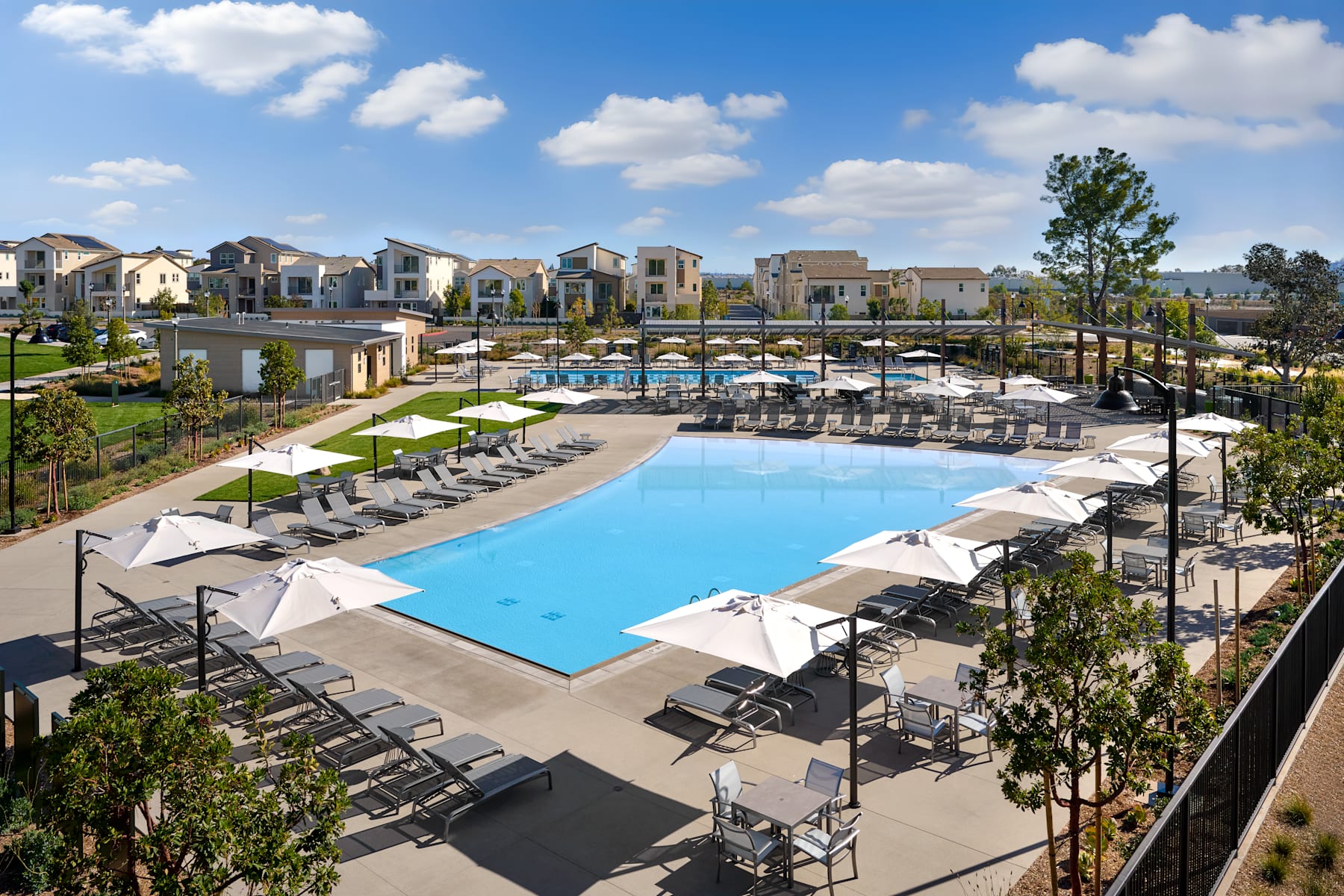 A large outdoor swimming pool surrounded by lounge chairs and umbrellas, with a residential community visible in the background under a blue sky with fluffy white clouds.
