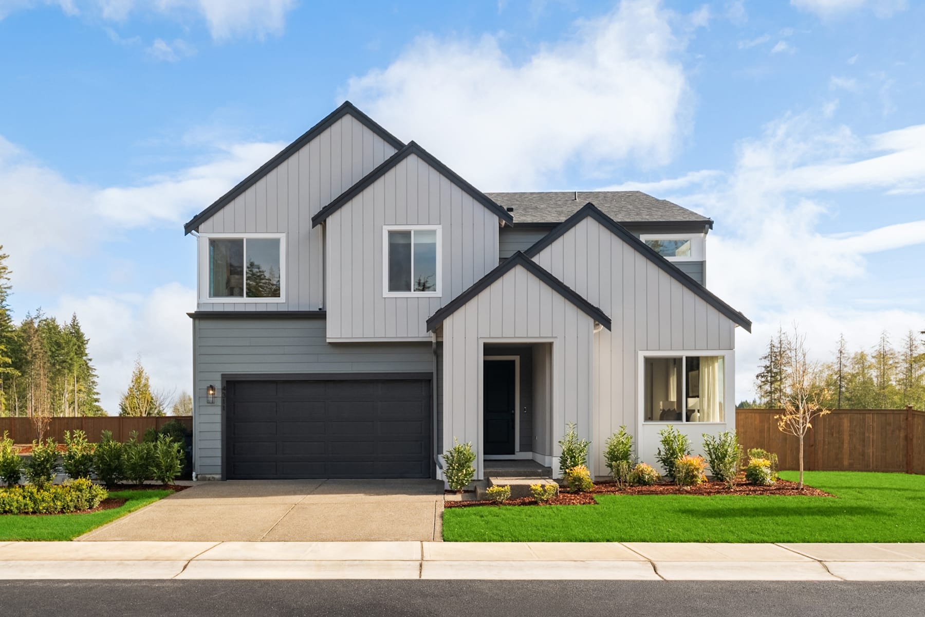 A modern two-story house with a gray exterior, a black garage door, and a well-manicured lawn with landscaping in the foreground, set against a backdrop of a blue sky with clouds.