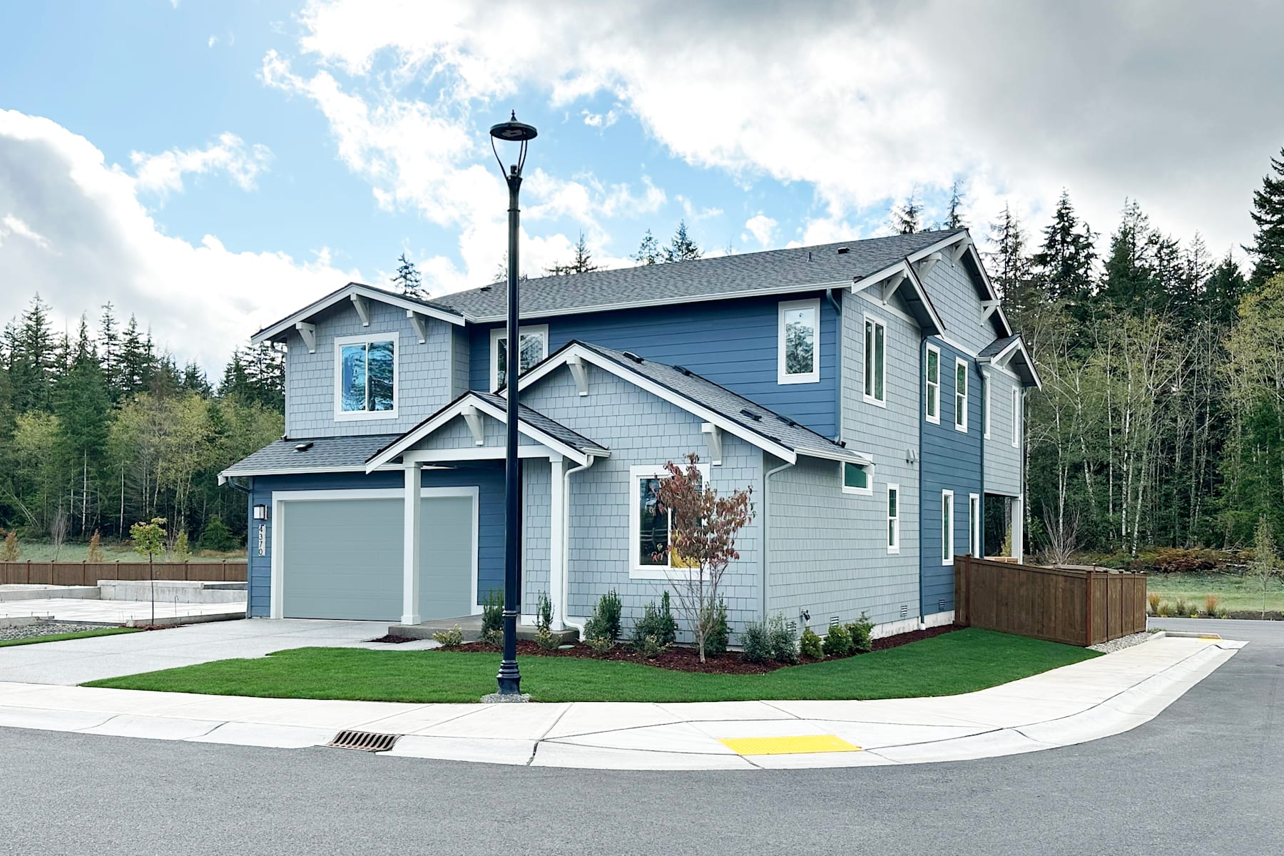 A two-story gray house with a garage, surrounded by a grassy lawn and trees, stands in a residential neighborhood under a partly cloudy sky.