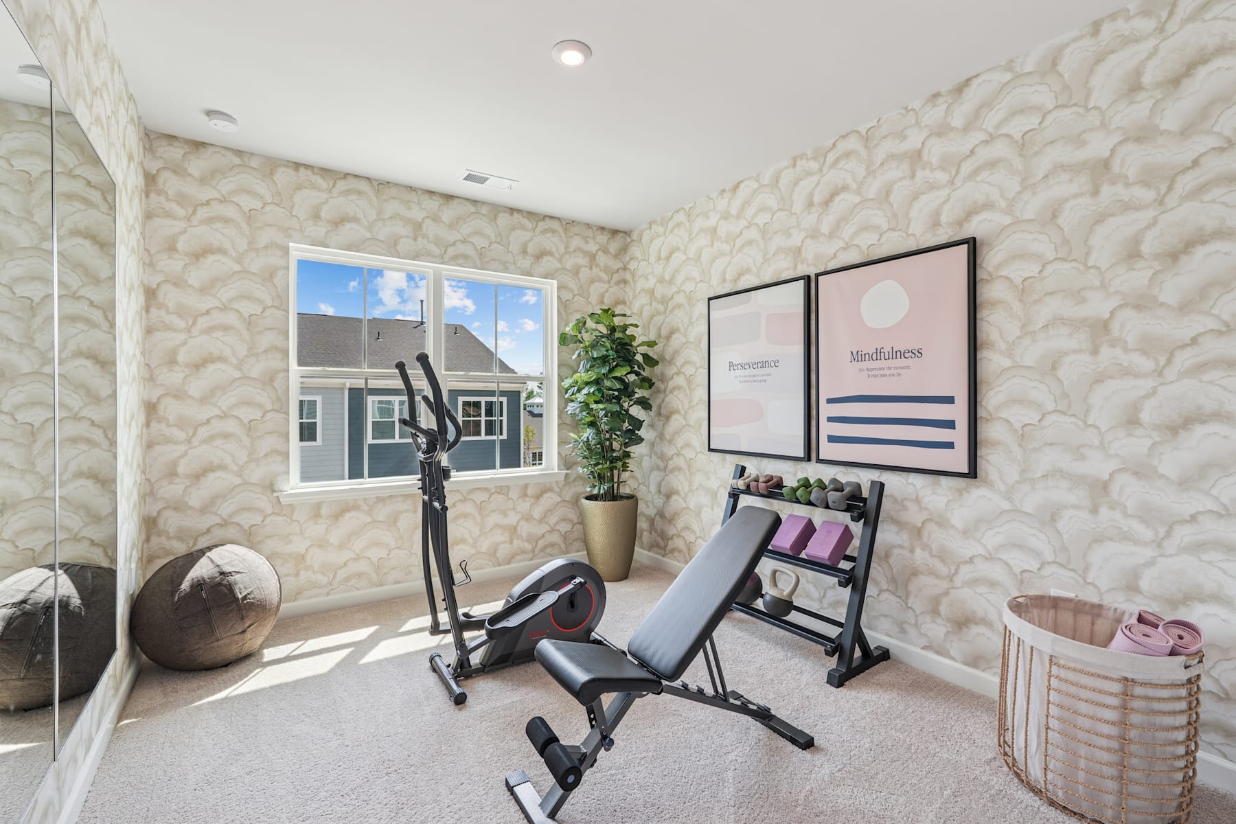 A cozy home gym with a variety of exercise equipment, including a weight bench and a stationary bike, set against a patterned wallpaper backdrop and large windows providing natural light.