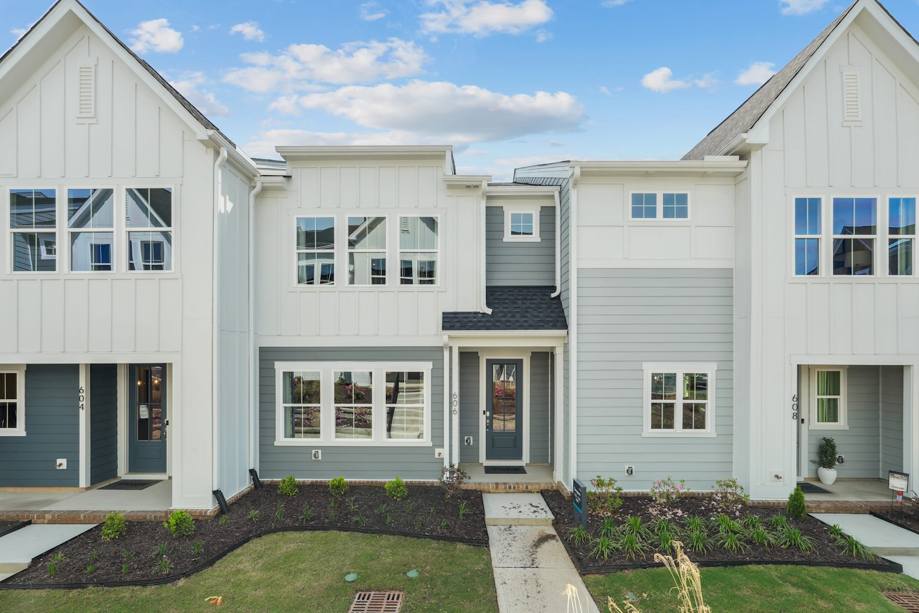 A modern, two-story townhouse with a well-maintained lawn and landscaping in the foreground, set against a backdrop of a clear blue sky with fluffy white clouds.