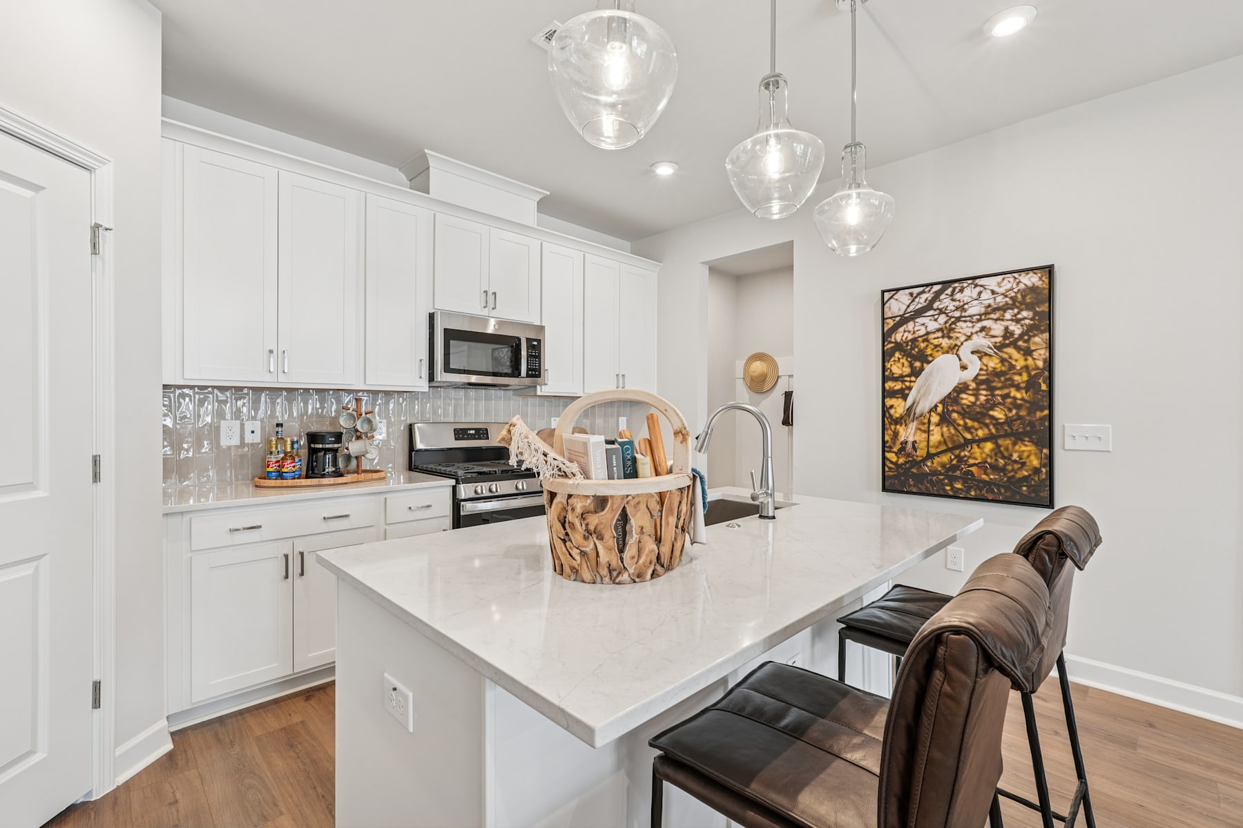 A modern, bright kitchen with white cabinets, a large island with a white countertop, and pendant lights hanging above. In the background, there is a framed artwork depicting a natural scene.