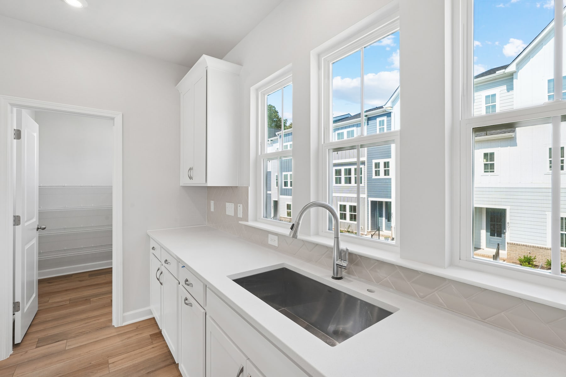 A modern, bright kitchen with large windows overlooking a cityscape, featuring white cabinets, a stainless steel sink, and hardwood floors.